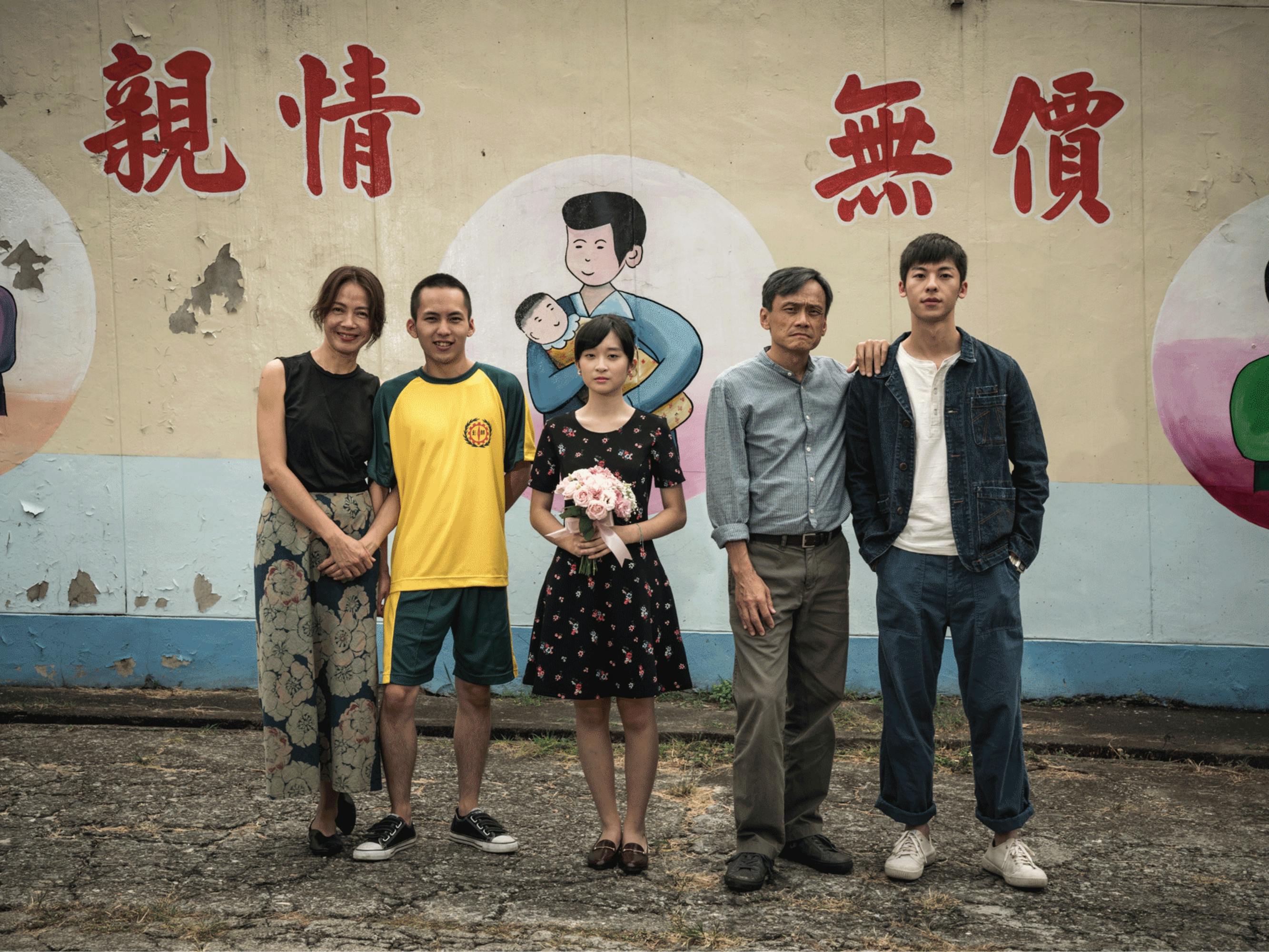 In this still from the film, the actors stand in front of a painted mural showing simple drawings of women holding infants. Wu Tai-ling, in the center, holds a bouquet of pink flowers. Samantha Ko and Wu Chien-ho stand in a pair on the left. Chen Yi-wen places a hand on Hsu Kuang-han’s shoulder.