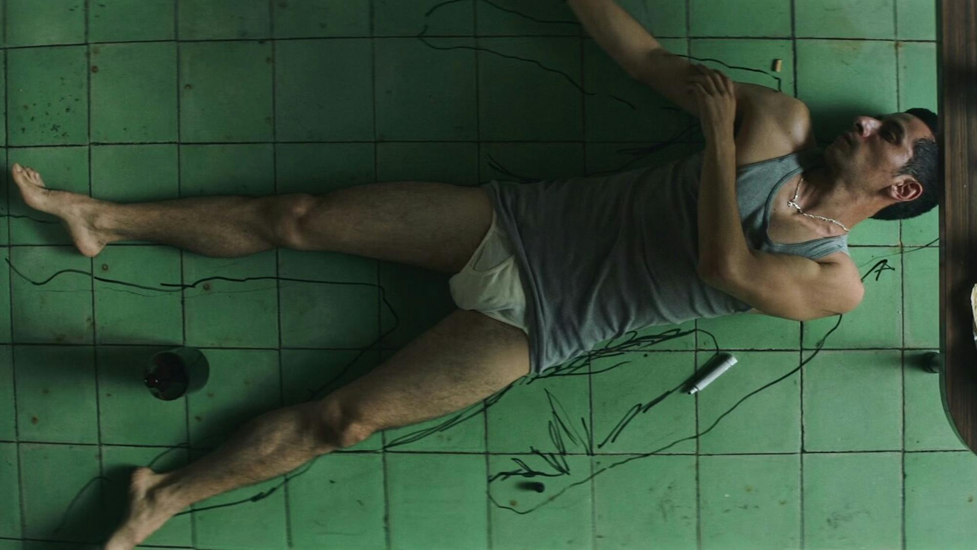 Raúl Briones wears a grey tank top and white underwear as he lies on a green tiled floor.