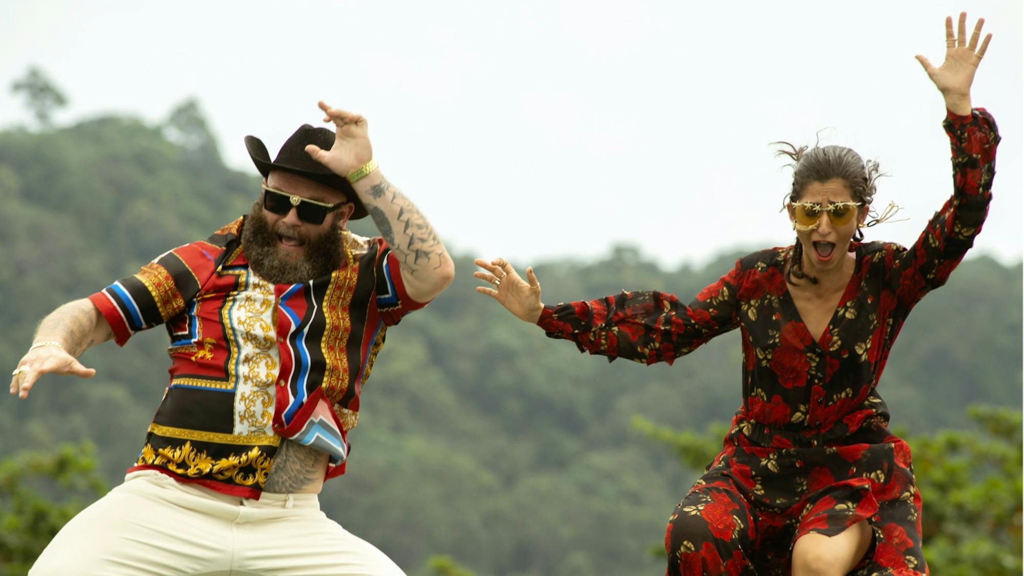 Helsinko (Darko Perić) and Nairobi (Alba Flores) jump, with alarmed expressions on their faces. Helsinko wears a colorful shirt, which reveals his tattooed body, sunglasses, and a cowboy hat. Nairobi wears a flowered dresses and glasses.