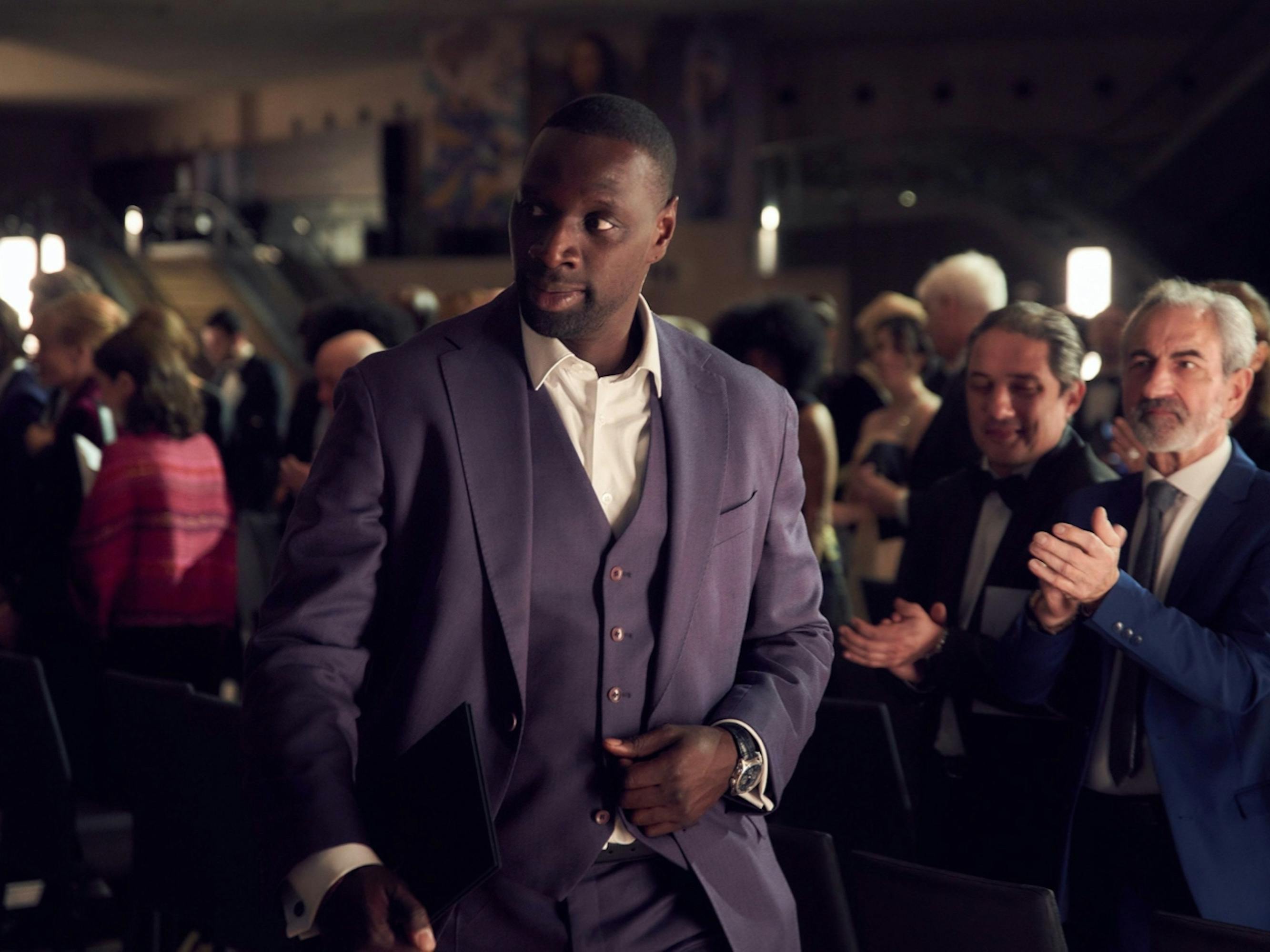 Assane Diop (Omar Sy) walks through a crowd as onlookers clap. He wears a stunning navy suit, watch, and white shirt.