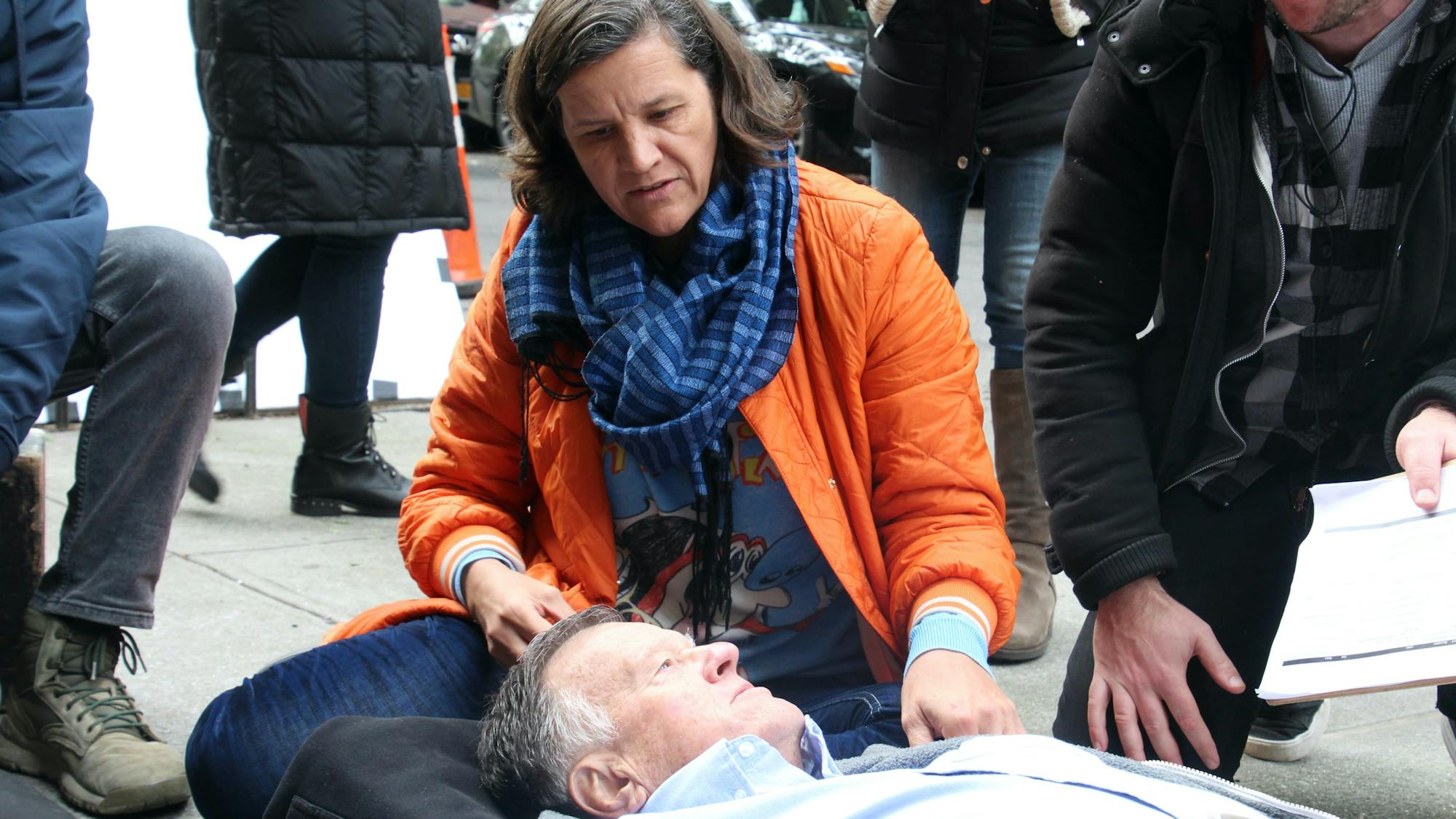 Kirsten Johnson directs her father in one of the deaths the film imagines for him. He lies on the sidewalk with his daughter and some of the crew flocking above him.