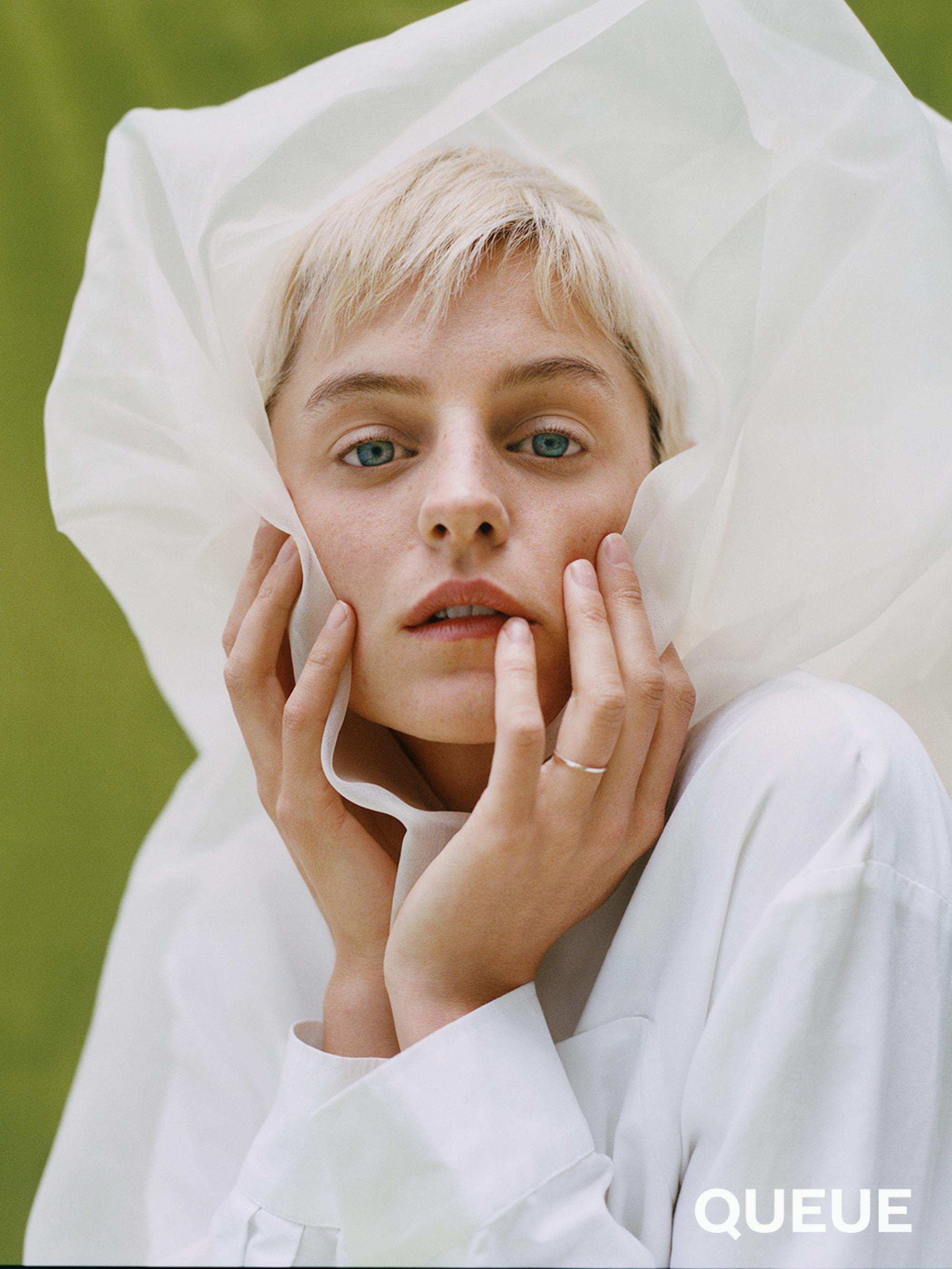 Emma Corrin wears a white top and a white hood and stands against a green drape.