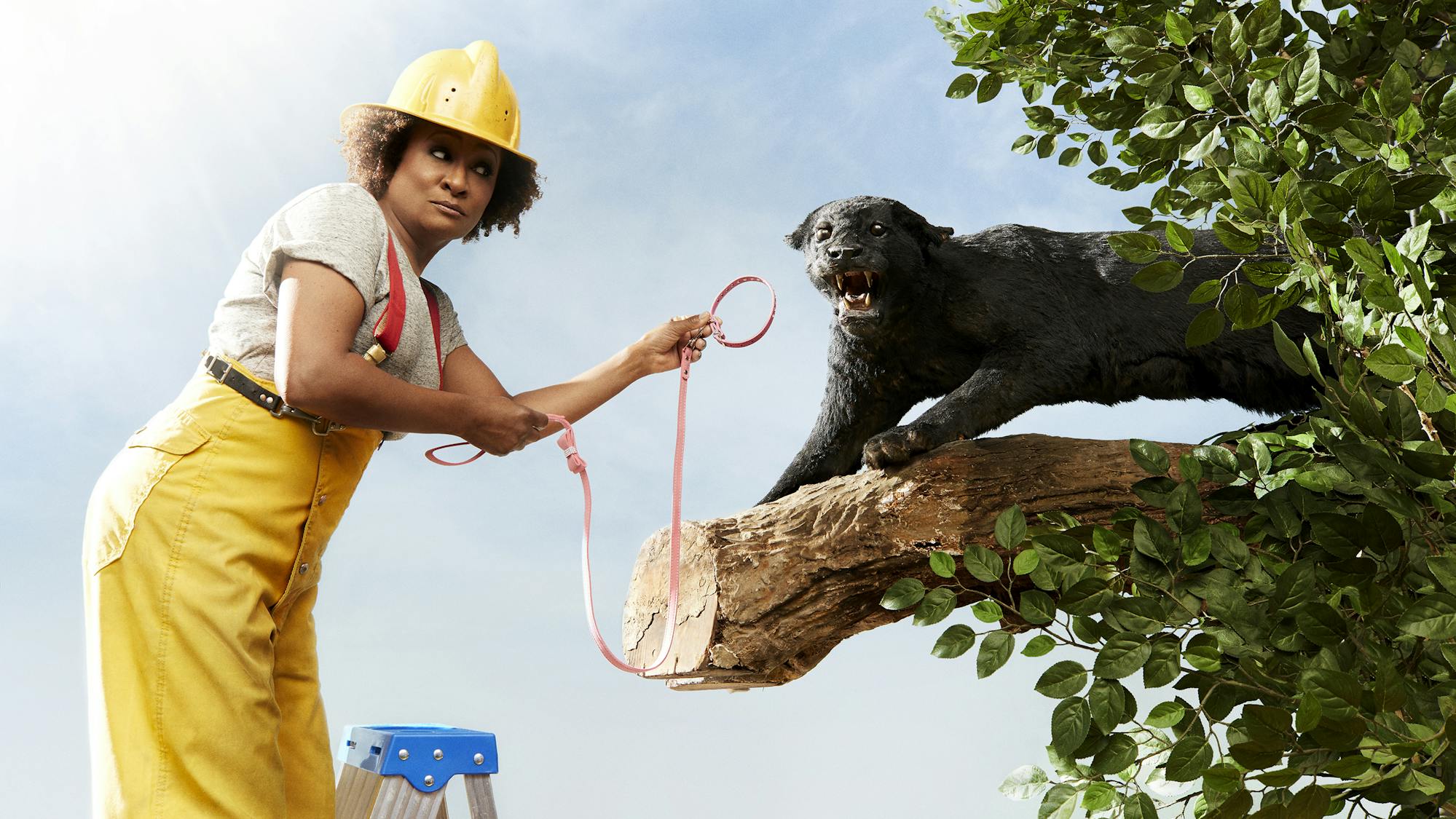 Wanda Sykes wears a yellow hard hat and yellow pants and holds a leash out to a taxidermied creatures.