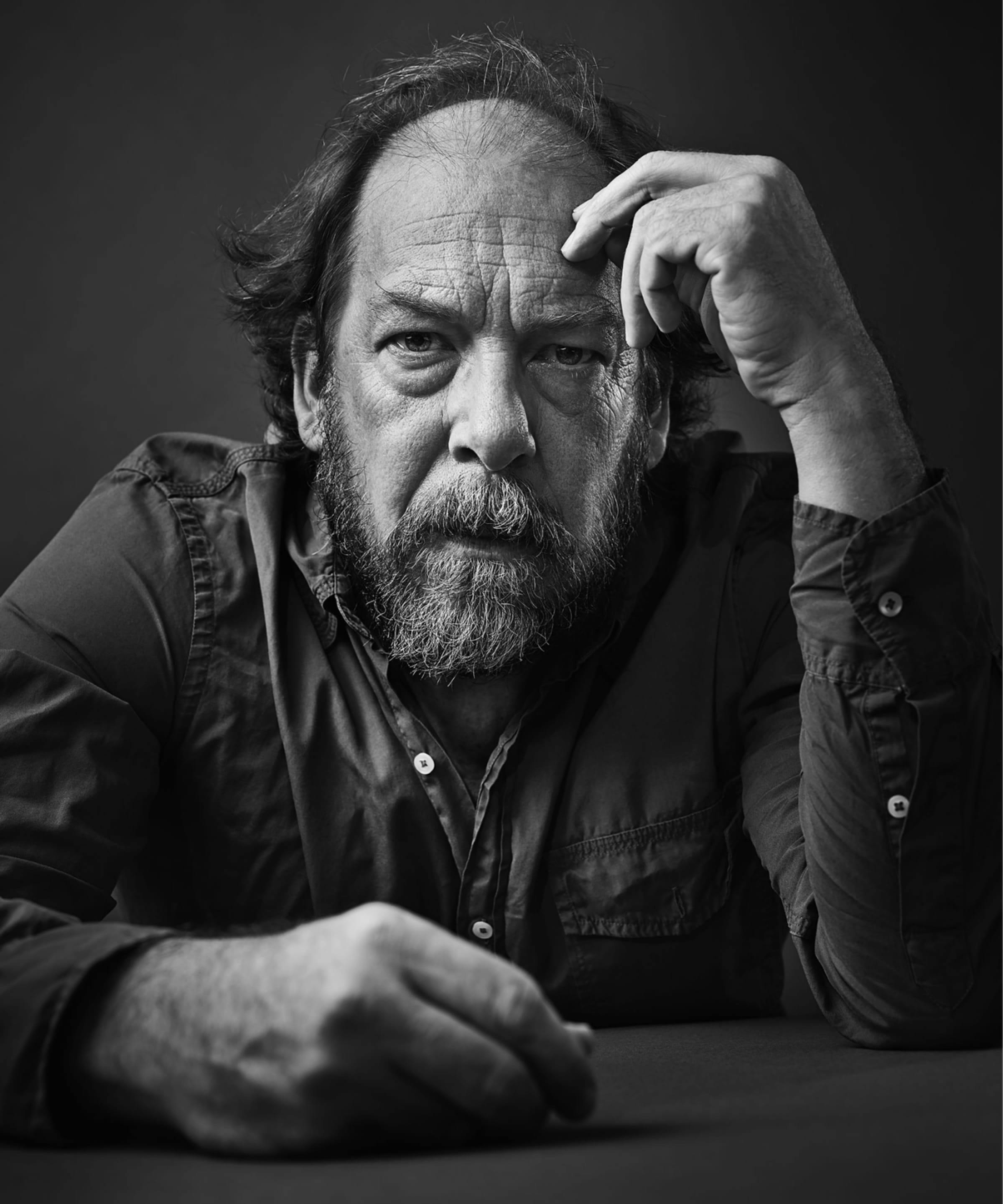 A black-and-white image of Bill Camp. He wears a dark buttoned down shirt, has a mustache and beard, and rests his fingers against his head. He looks very seriously right at the camera. 