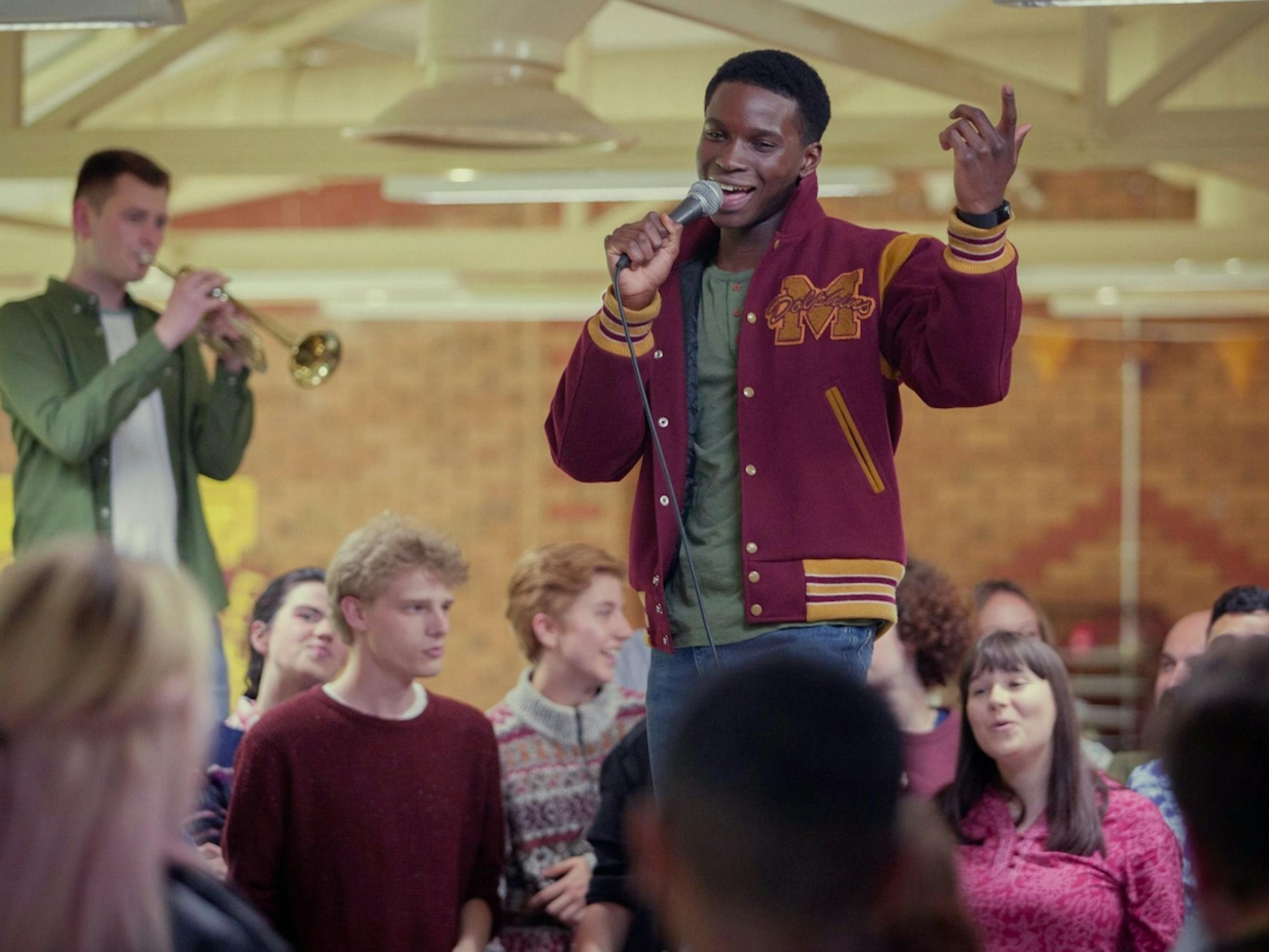 Jackson Marchetti stands around a bunch of students, flanked by members of the bank. He wears his letterman jacket and sings into a mike.