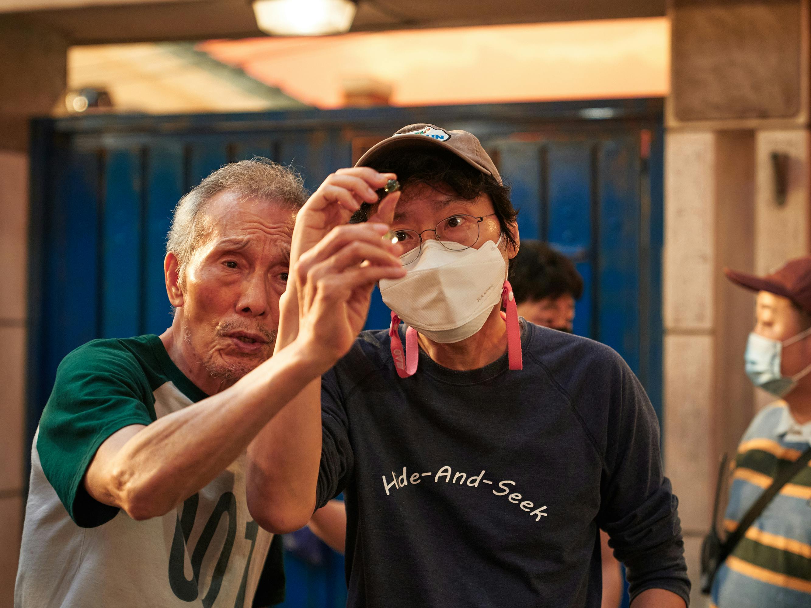 Oh Young-soo and Hwang Dong-hyuk look at a marble. Young-soo wears a white and green baseball style shirt. Director Hwang wears a 'hide-and-seek- black t-shirt, baseball hat, and white mask.