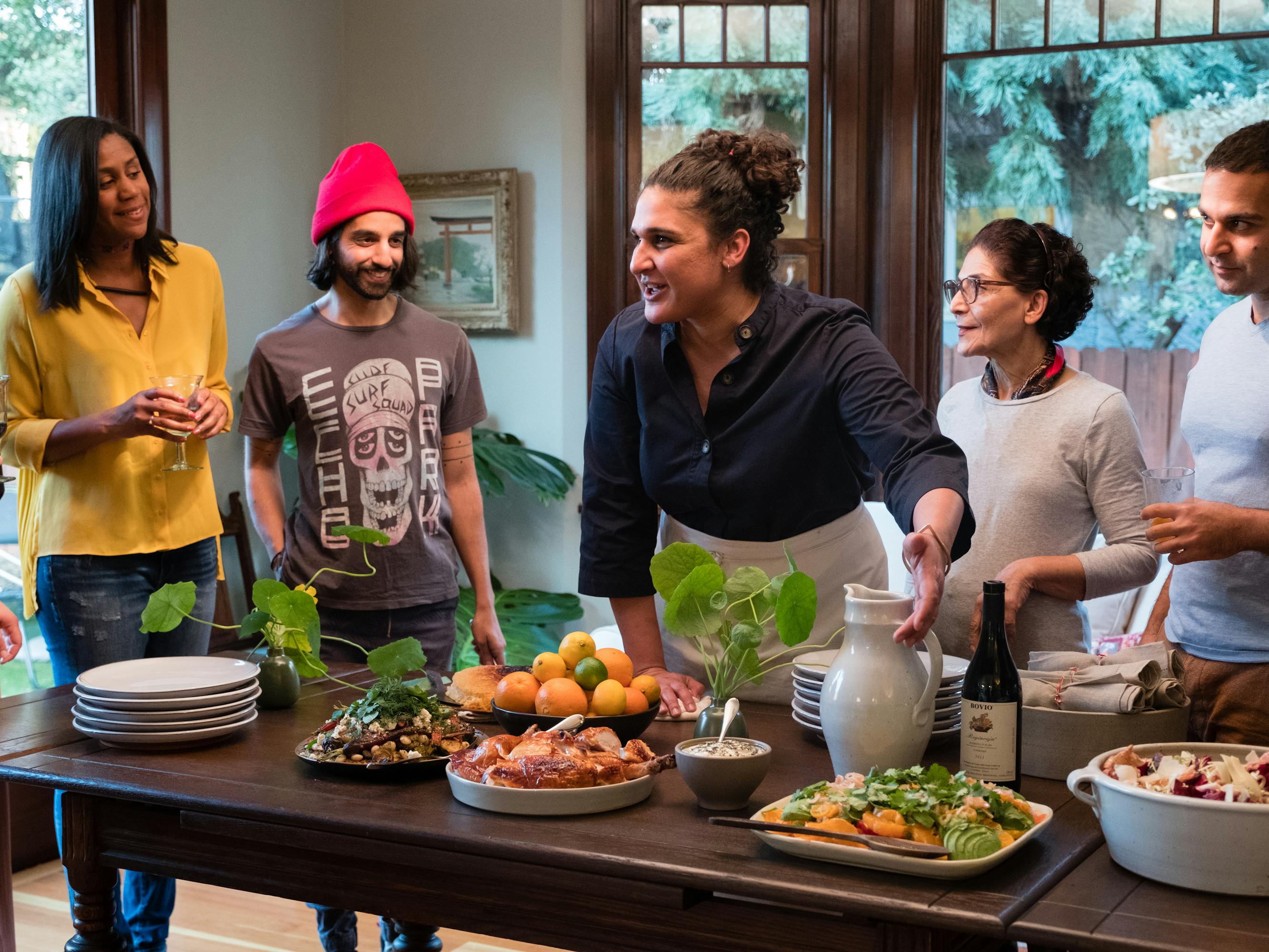 Samin Nosrat stands around a table with five others gesturing to some of the dishes.