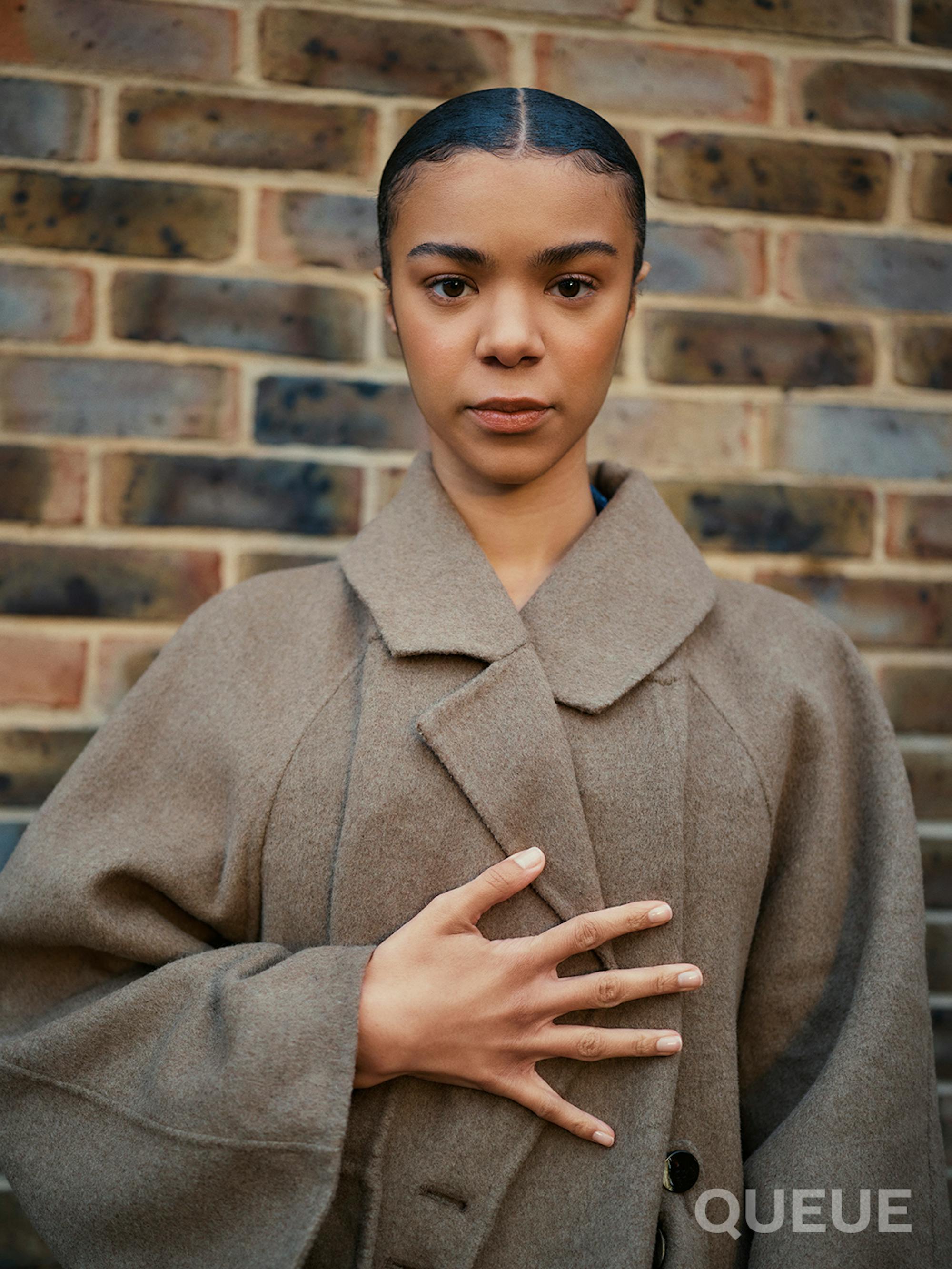 India Amarteifio wears a brown coat and stands against a brick wall. She closes the coat closed with her outstretched palm.