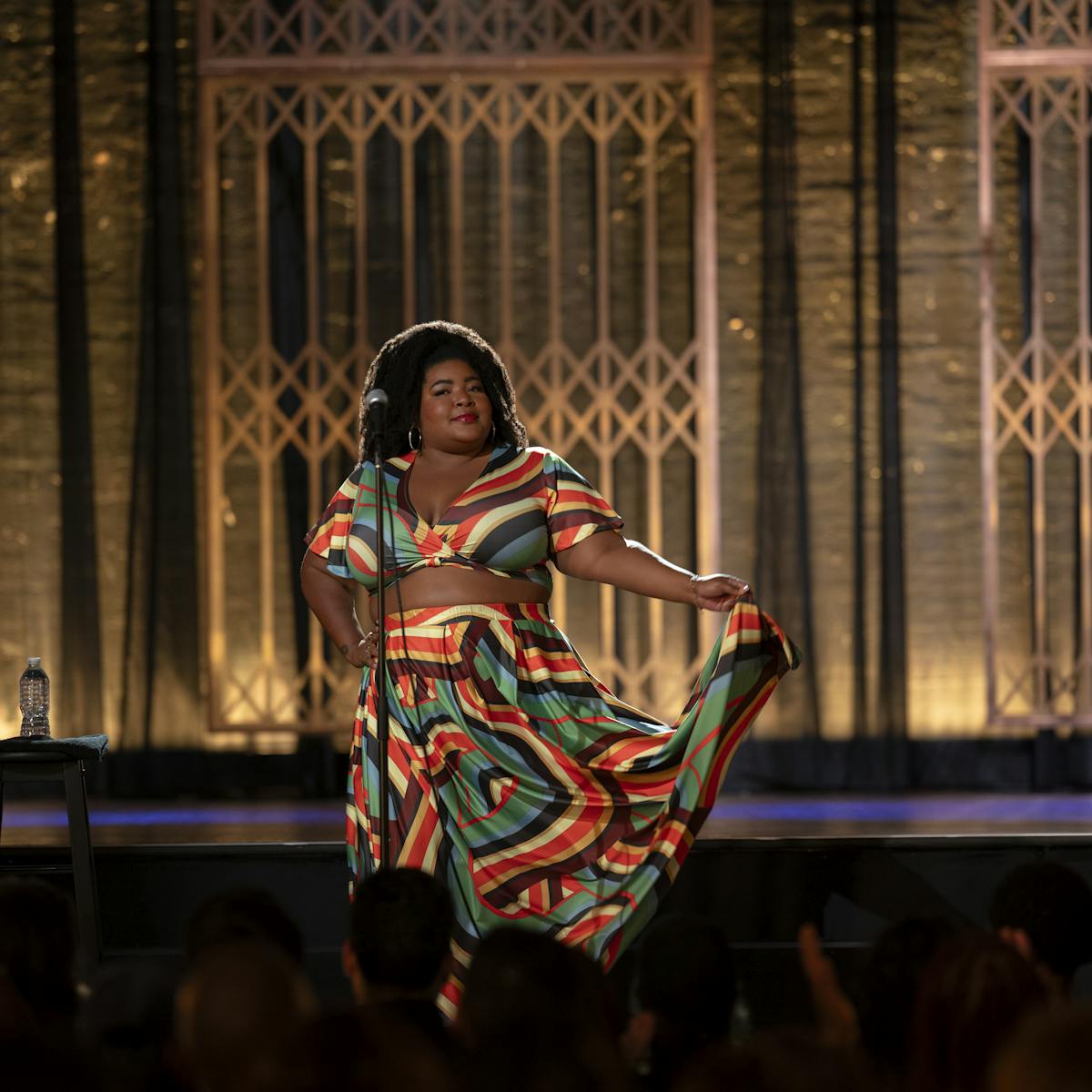 Dulcé Sloan wears a patterned skirt-and-top set and stands on an art-deco stage.