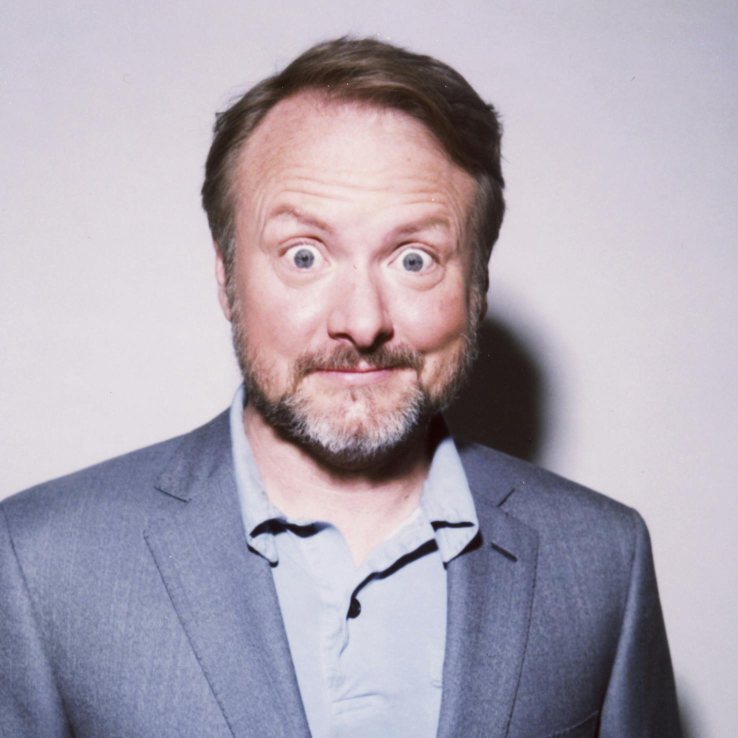 Rian Johnson wears a grey polo shirt and dark grey blazer and makes a silly face at the camera.