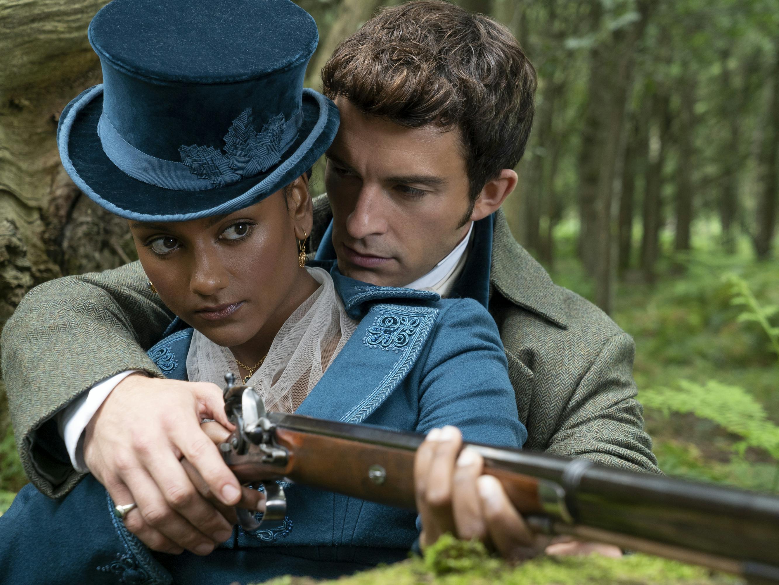 Kate Sharma (Simone Ashley) holds a gun and Anthony Bridgerton (Jonathan Bailey) wraps his arms around her with instruction. 