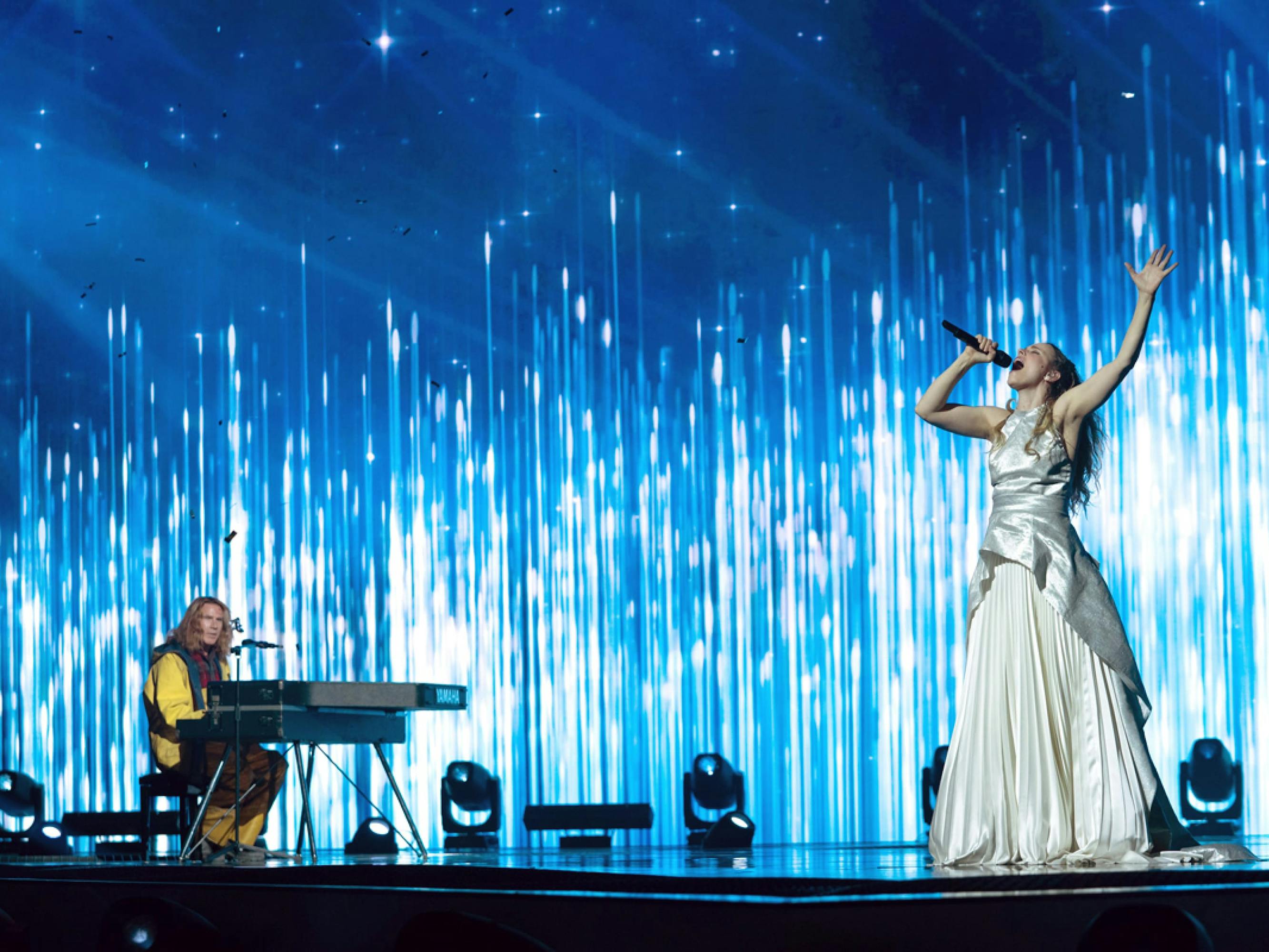 Will Ferrell sits behind a keyboard on a stage, wearing a yellow fishing outfit. Rachael McAdams stands closer to the edge of the stage, singing with her arm raised up as she belts out the song “Húsavík” in a white and silver gown.