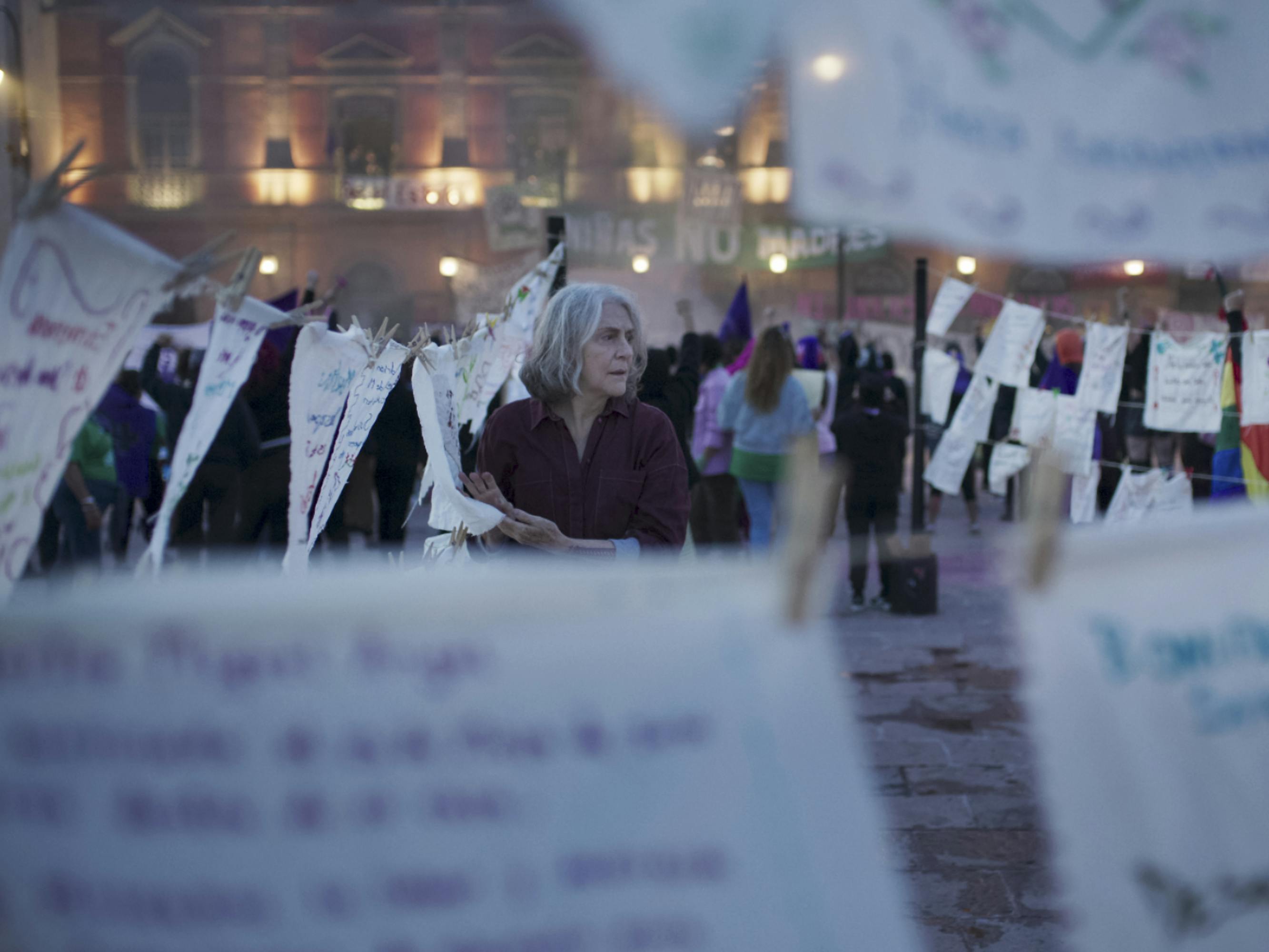 Una mujer se encuentra entre hojas de papel blanco colgadas en tendederos. Detrás de ella hay un enorme edificio débilmente iluminado por luces individuales en cada ventana.