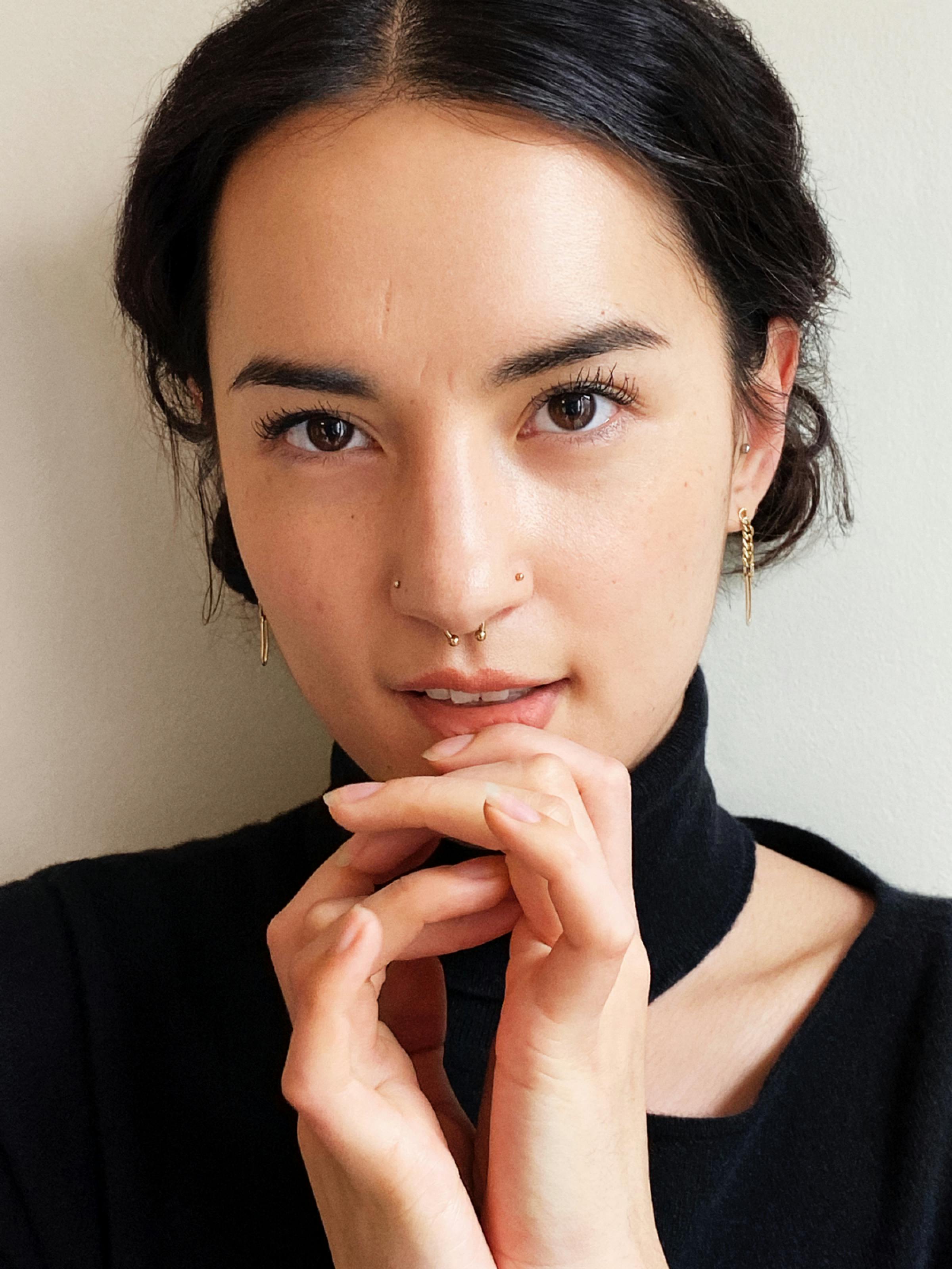 Jessie Mei Li wears a black turtleneck baring a strip of skin. She wears gold earrings, a few nose rings, and holds her hands beneath her mouth.