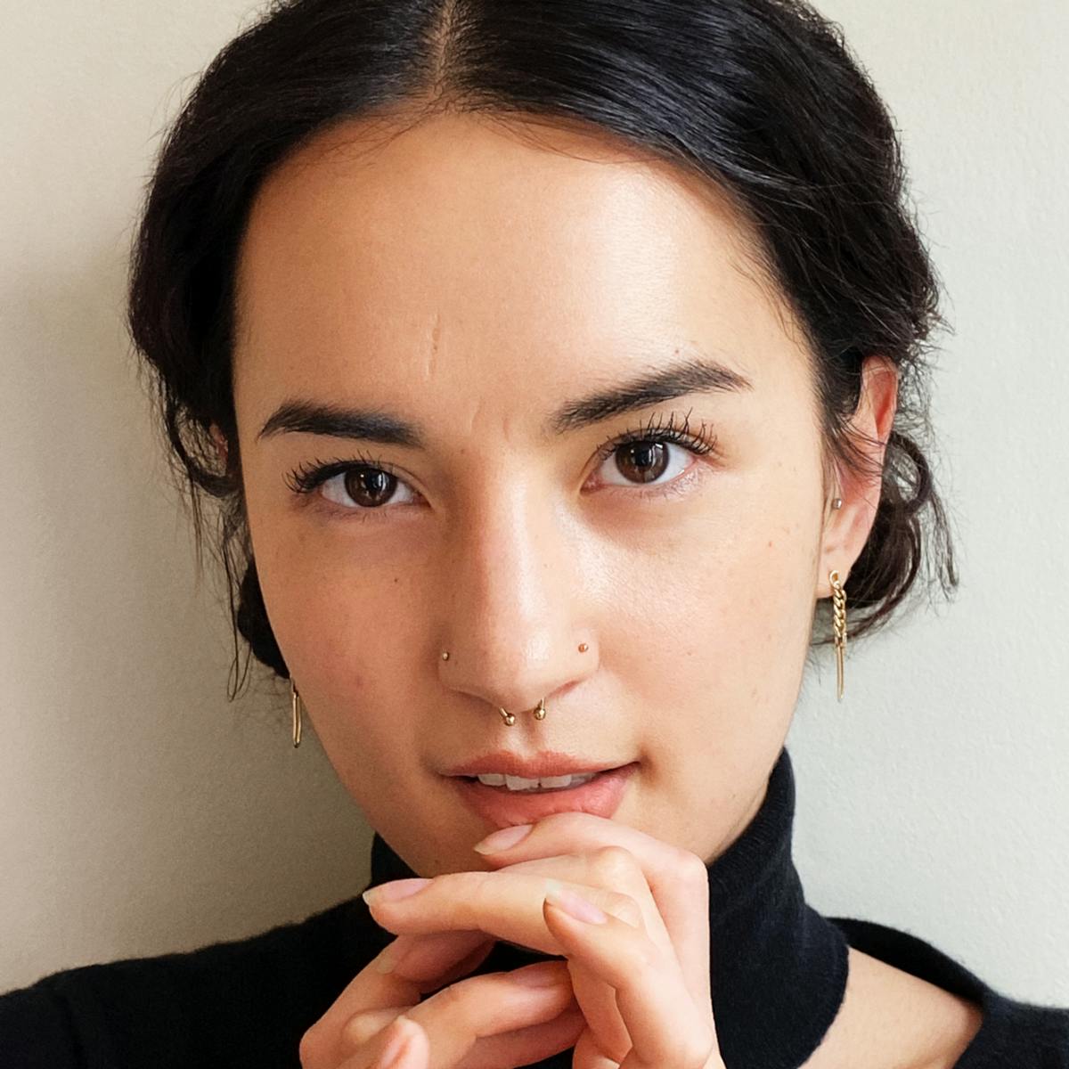 Jessie Mei Li wears a black turtleneck baring a strip of skin. She wears gold earrings, a few nose rings, and holds her hands beneath her mouth.