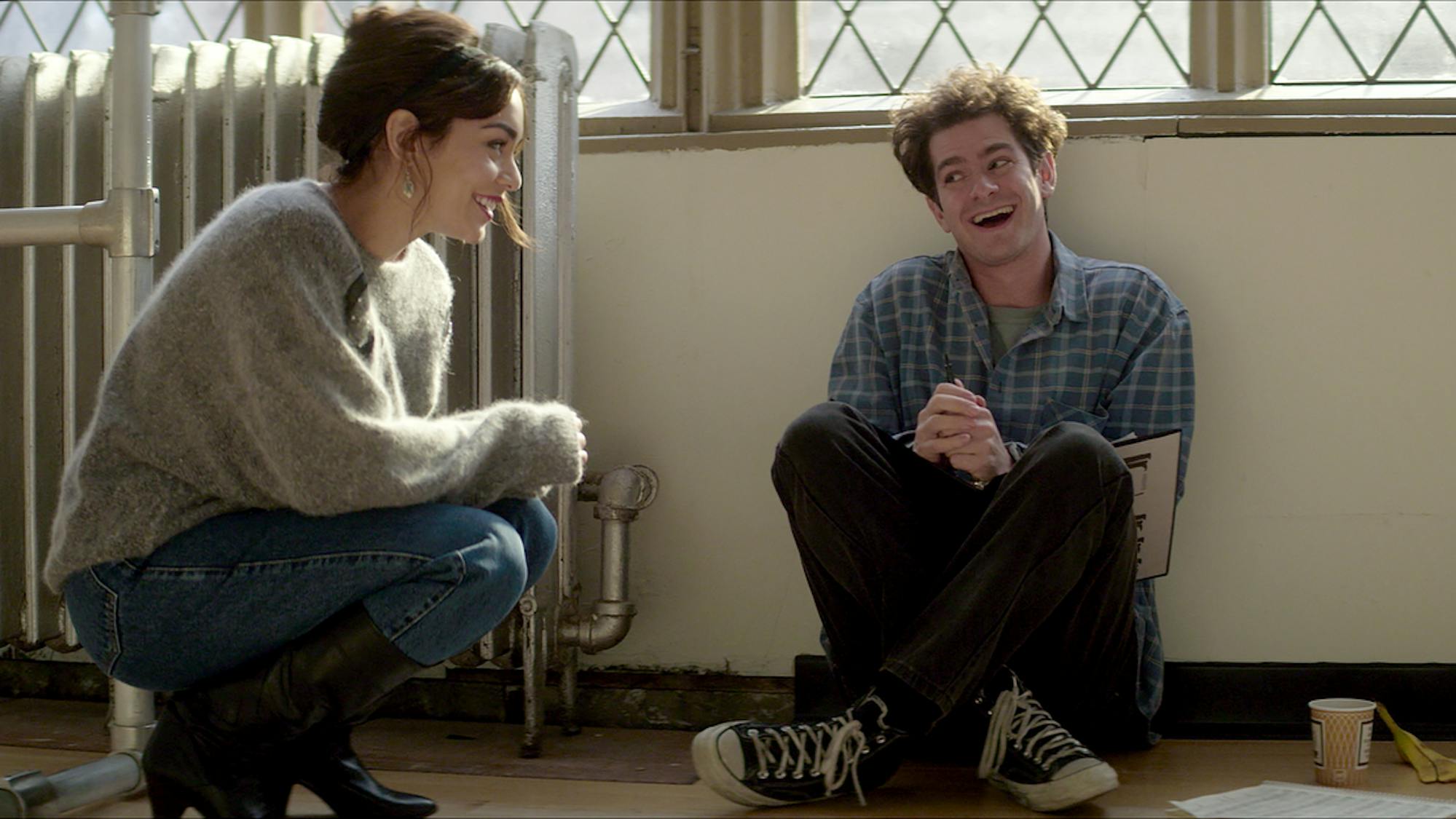 Vanessa Hudgens and Andrew Garfield as Jonathan Larson sit in a rehearsal space. Hudgens wears jeans and a light colored sweater. Garfield wears converse, dark pants, and a checkered shirt. Light streams in from the windows behind them.