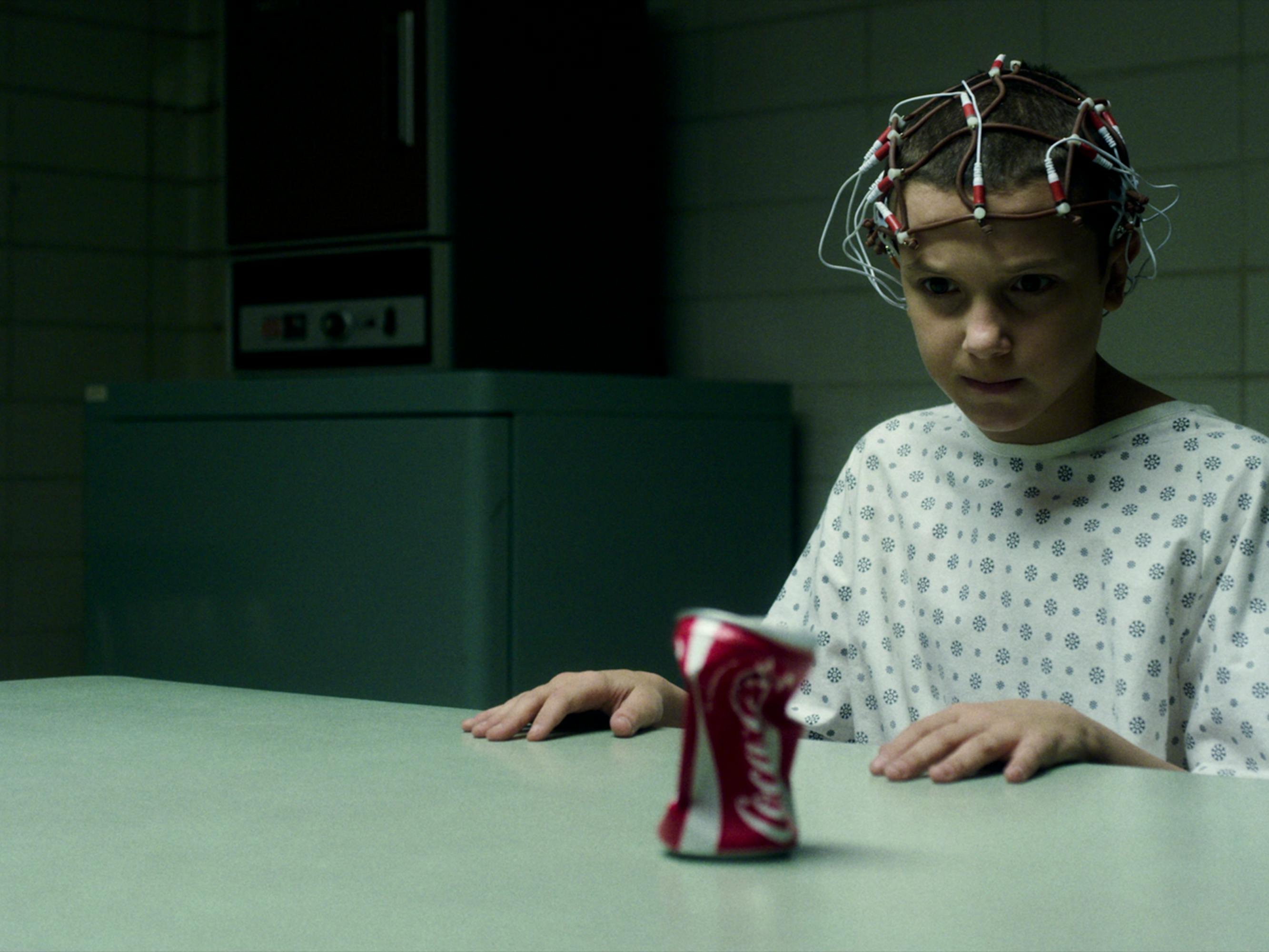 Eleven (Millie Bobby Brown) wears a hospital gown with wires on her head. She stares at a crumpled can of coke.