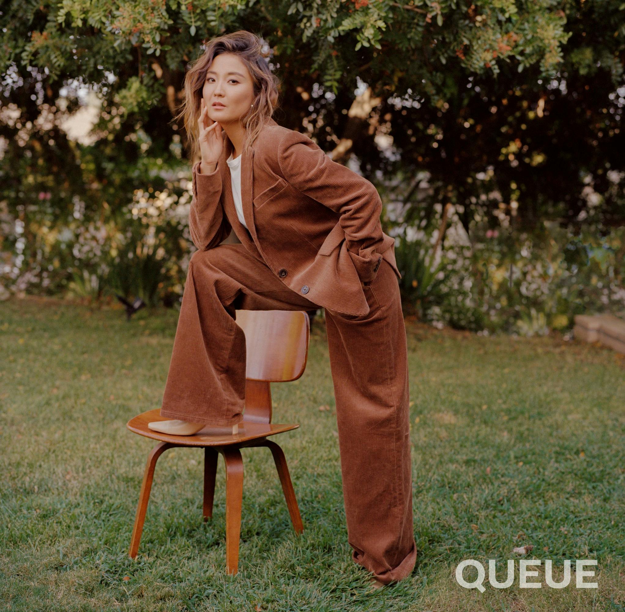 Ashley Park wears an oversized brown suit. She stands with one leg planted on green, sunburnt grass, and the other bent on a wooden chair. She looks very chic and powerful.