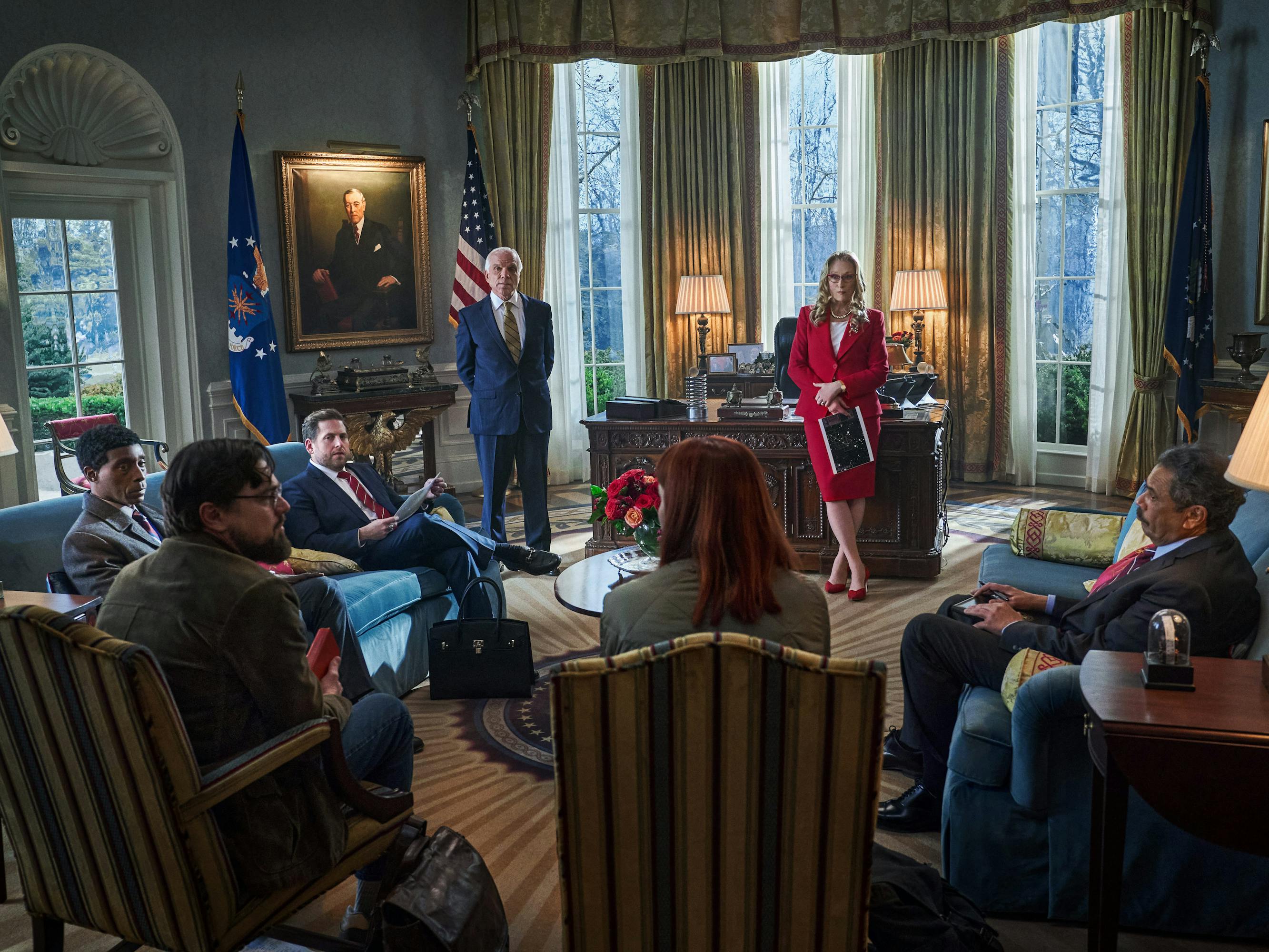 Rob Morgan, Leonardo DiCaprio, Jonah Hill, Jennifer Lawrence, and Meryl Streep sit in the oval office. Morgan wears a grey suit. DiCaprio wears a green corduroy jacket. Hill wears a navy suit with a black Birkin at his feet. Lawrence wears a green top, but only her bright red hair is visible. Streep wears a fabulous red skirt suit with matching heels. 