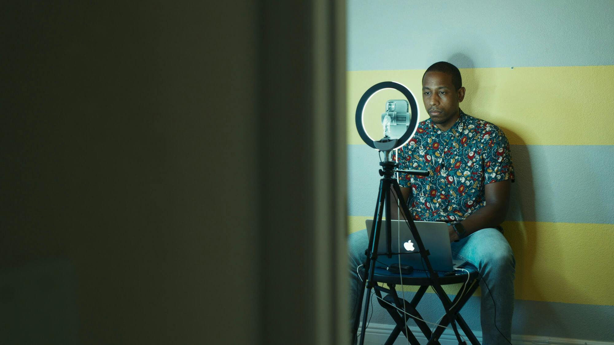 Dr. Armen Henderson sits in front of a computer and ring light. He wears jeans and a patterned shirt. The wall behind him is green and yellow striped.