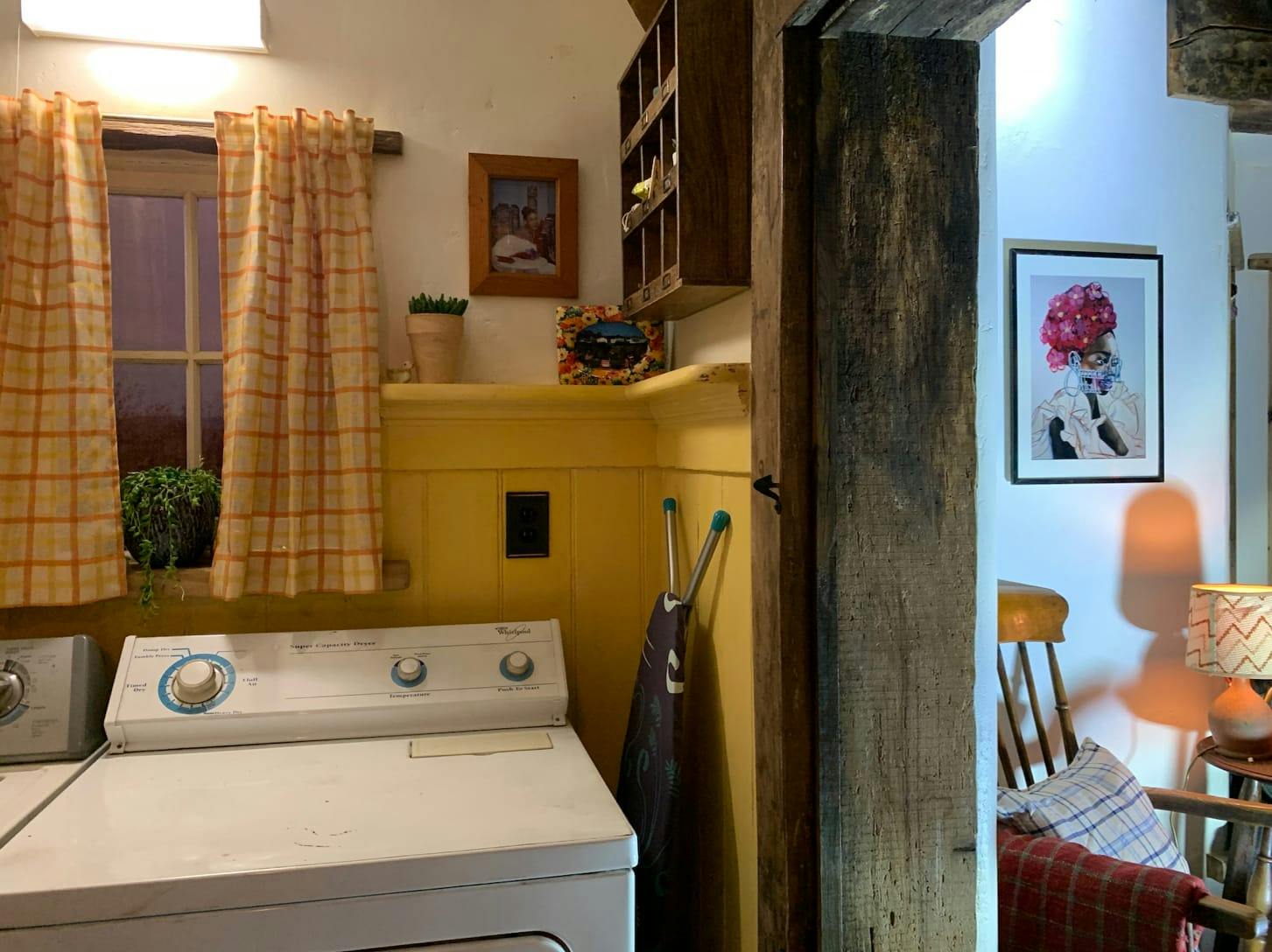 The laundry room, which is finished with a bright yellow. The washer and dryer are white, there is an ironing board, checkered curtains, and a wood panel.