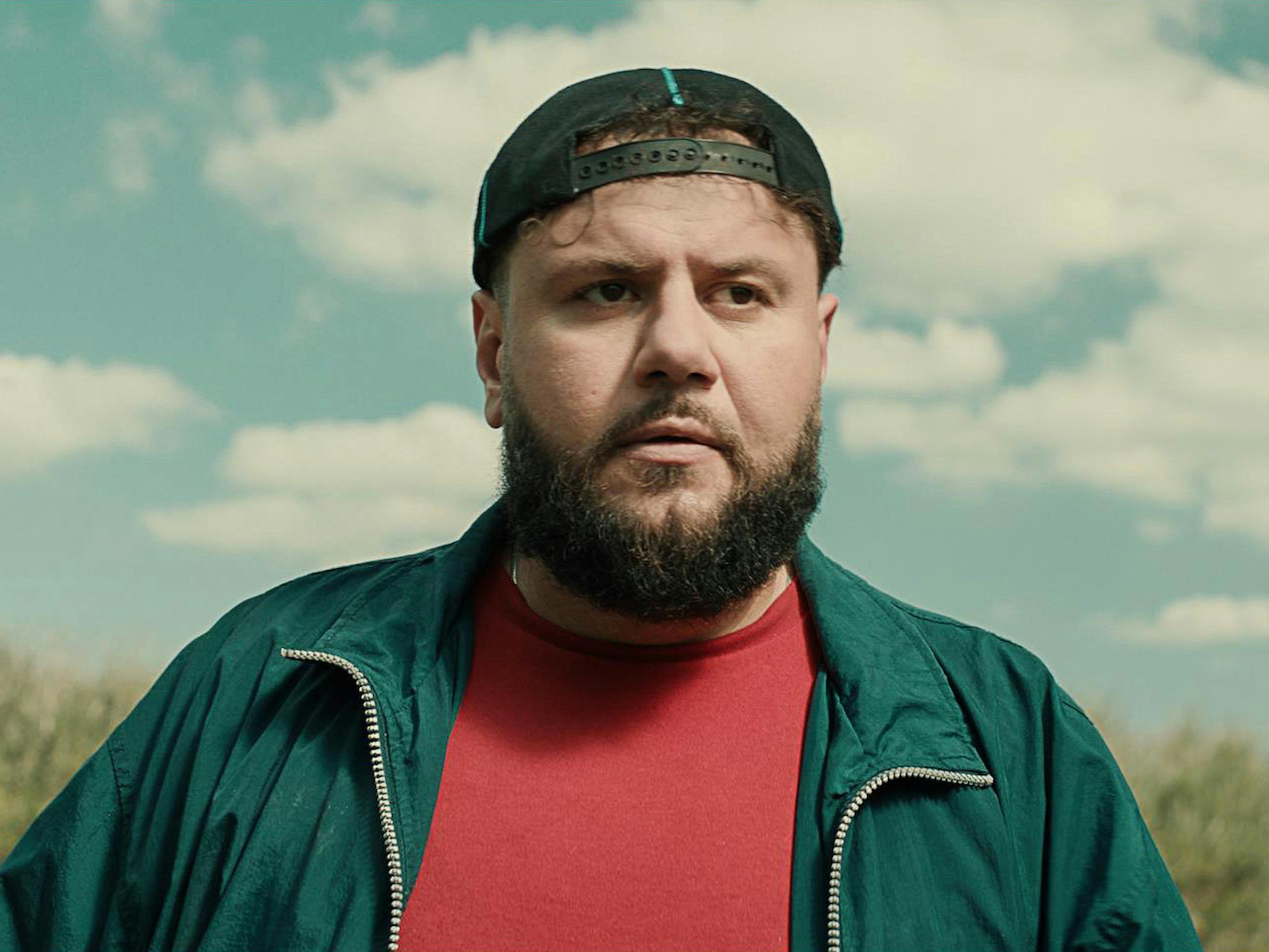 Mo (Mo Amer) wears a red shirt and green jacket, and wears a backwards hat. Behind him in a blue sky streaked with clouds.