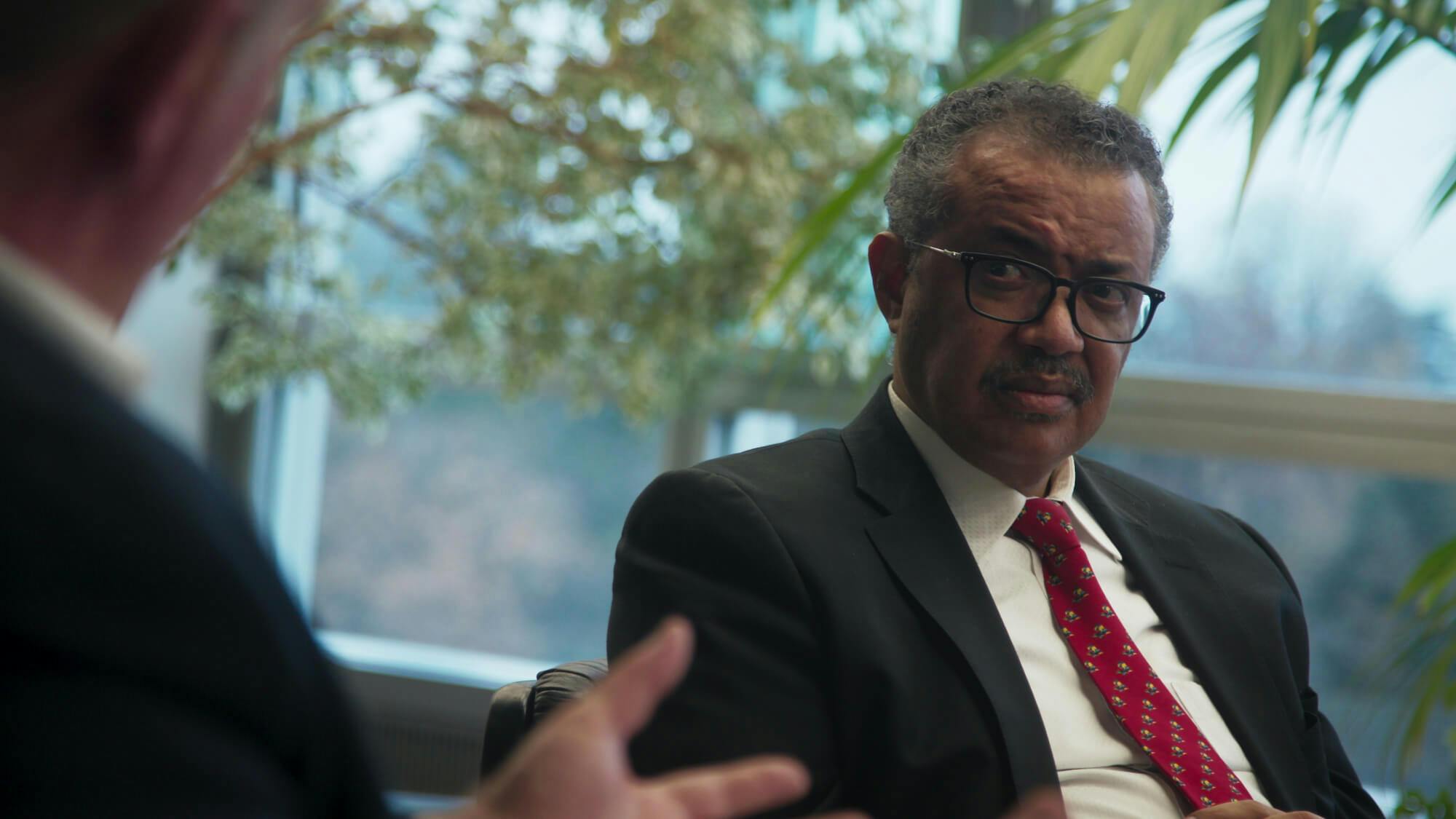 Dr. Tedros Adhanom Ghebreyesus speaks to someone off-camera and wears a dark suit, white shirt, glasses and red tie.