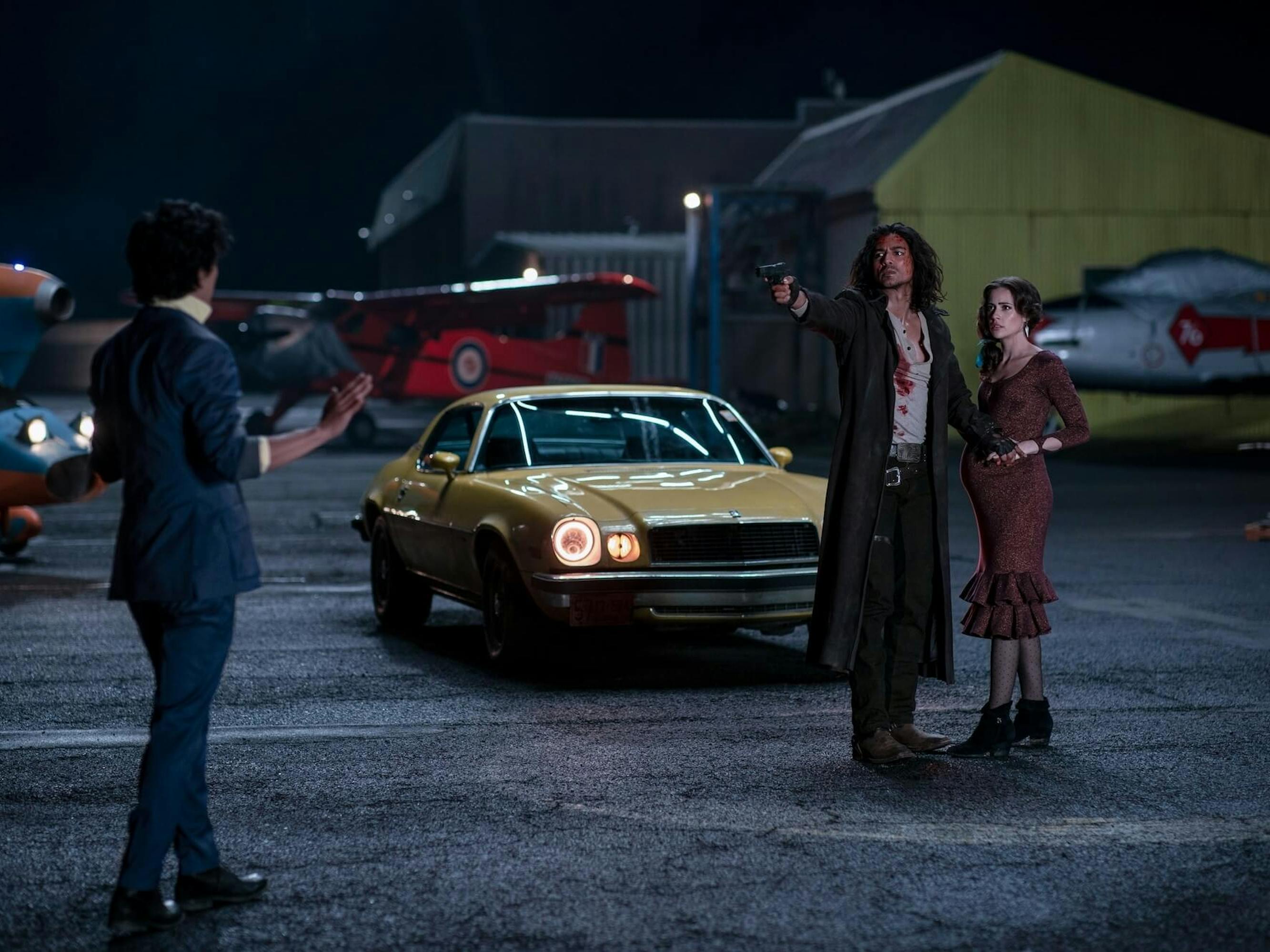 John Cho, Jan Uddin, and Lydia Peckham face off in a parkinglot filled with planes. Uddin points a gun at Cho as he shields a pregnant Peckham. Behind the duo is a yellow car.