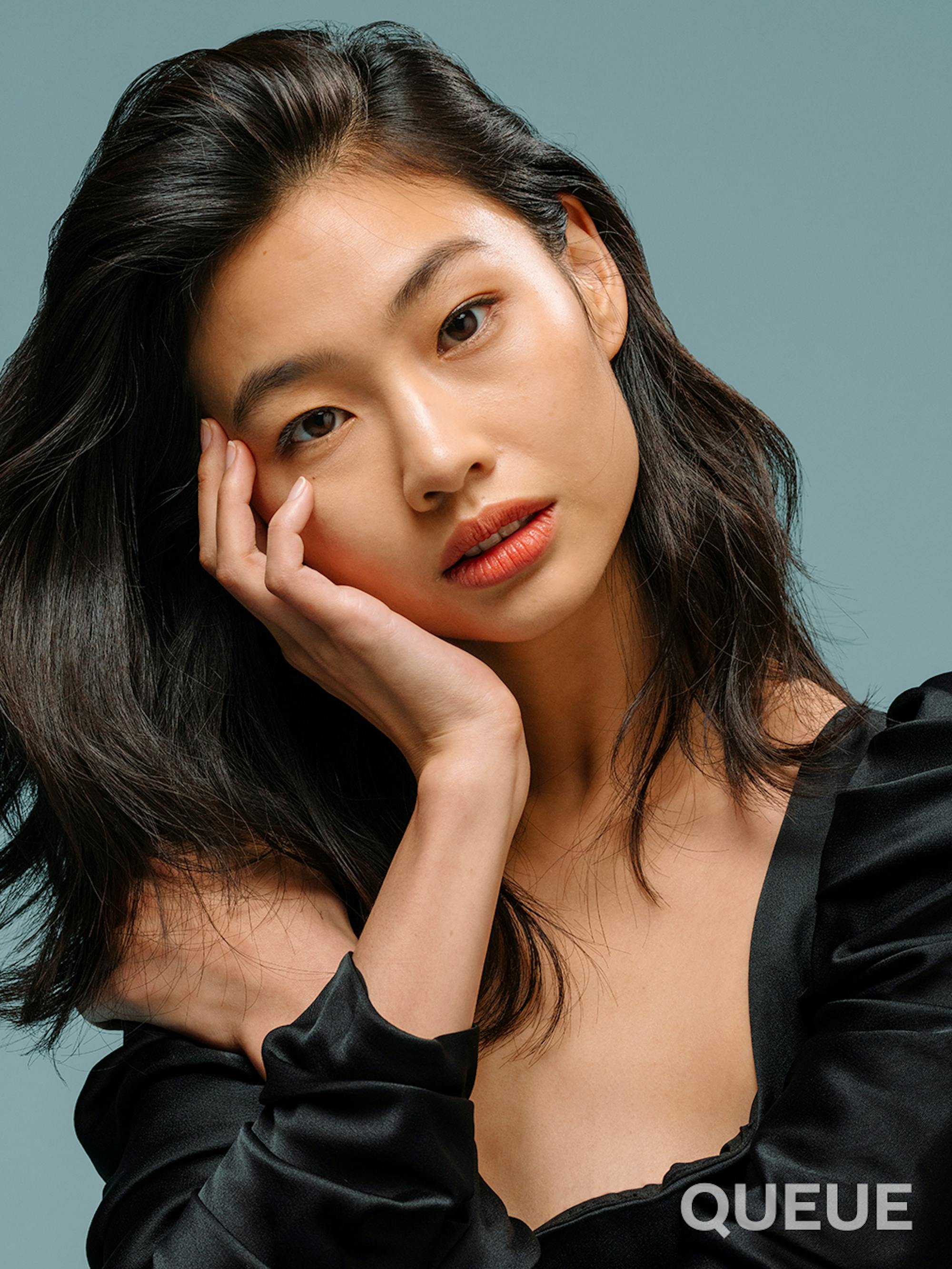 Jung Ho-yeon in a tight crop with her head tilted and one hand resting on her face, the other behind it placed on her shoulder and her hair flows to the left. She is wearing a satin black dress.