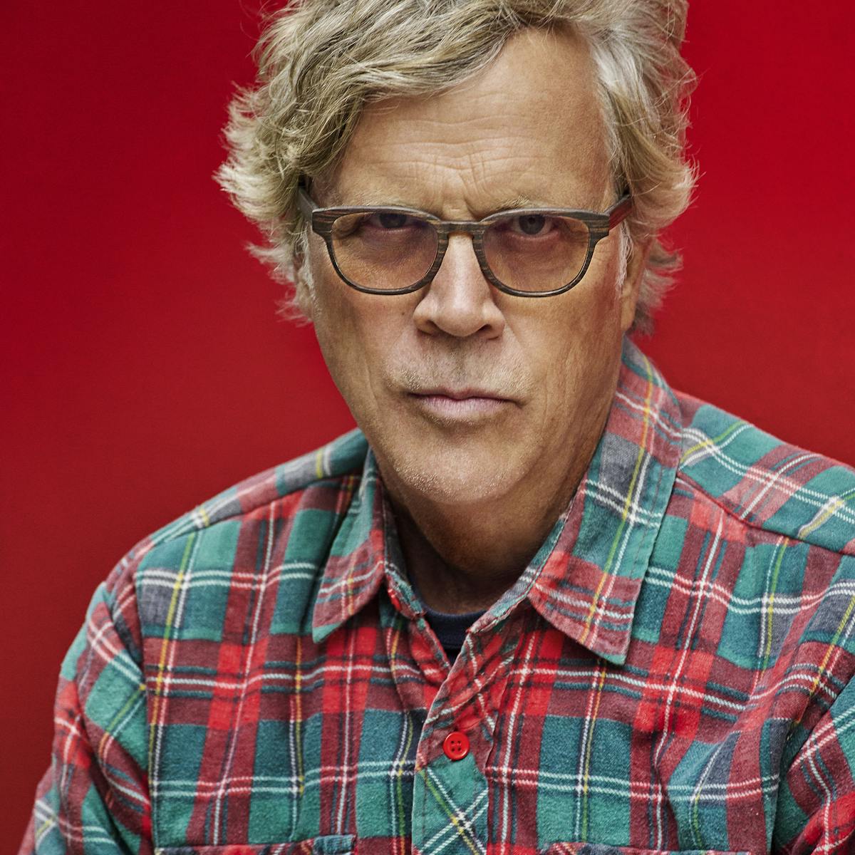 Todd Haynes wears a Christmas-y plaid flannel shirt and dark glasses against a red background.