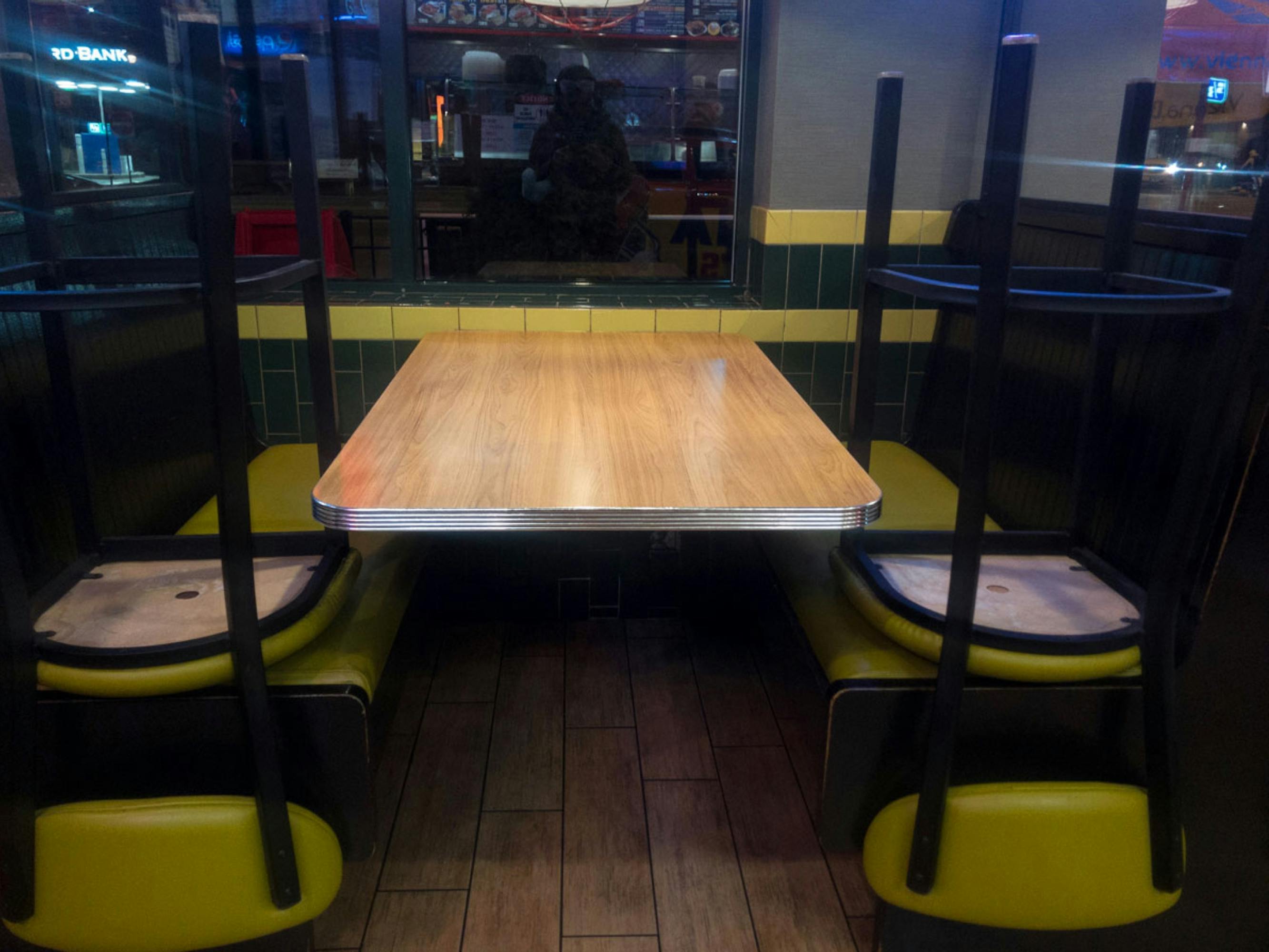 A photograph of an empty, green and yellow restaurant booth, charis set upside down as if at closing time. There is a neon glow to the picture, and you can see the reflection of the photographer in the window.