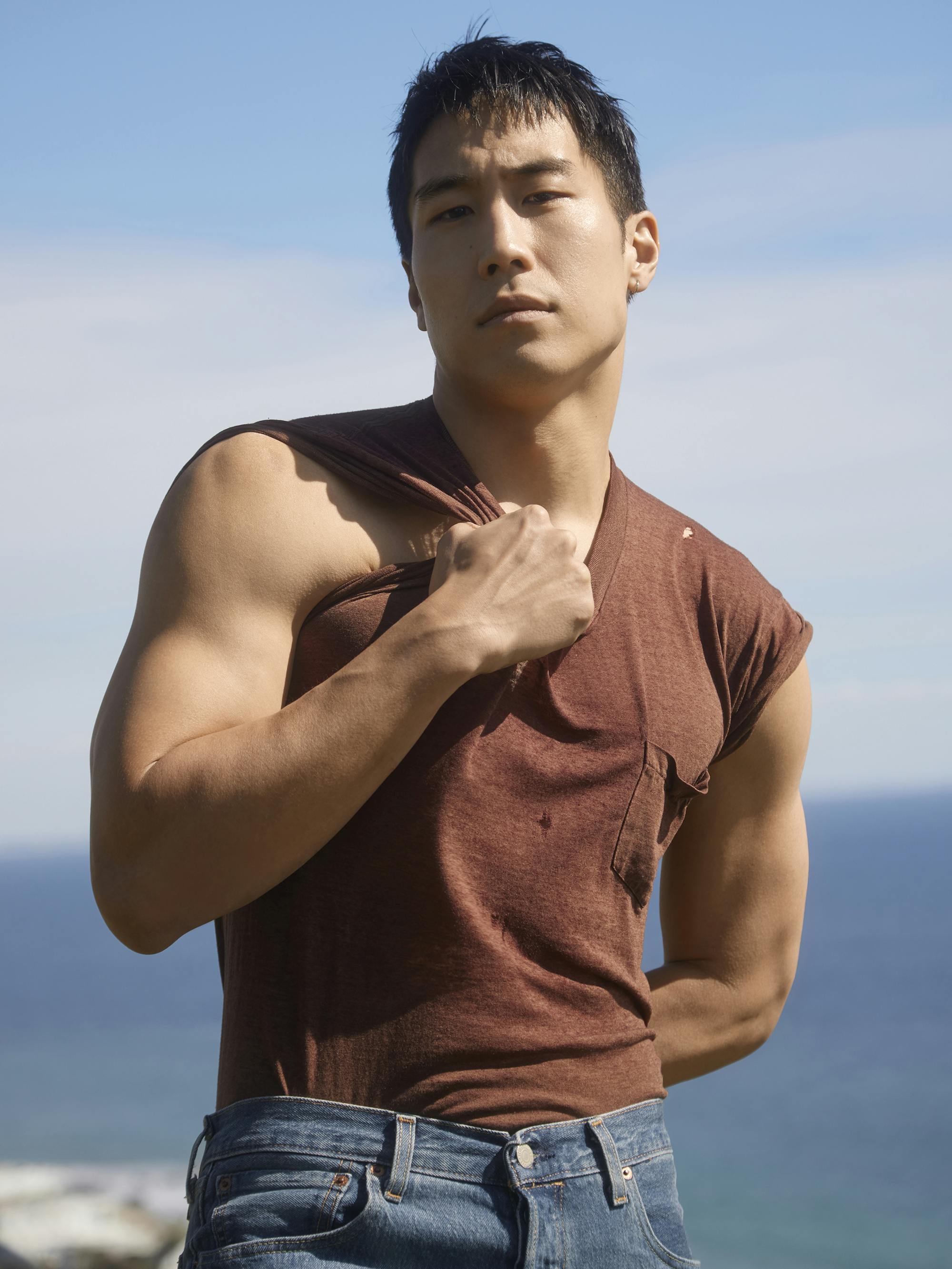 Young Mazino wears jeans and a brown tank top on a ledge overlooking the ocean. 