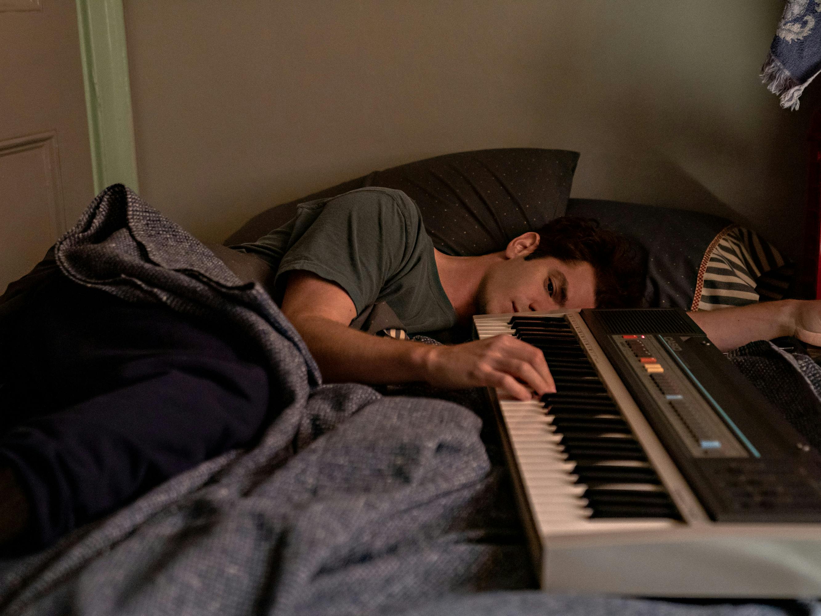 Andrew Garfield plays a keyboard piano in his bed. He looks forlorn as he rests his fingers against the keys.