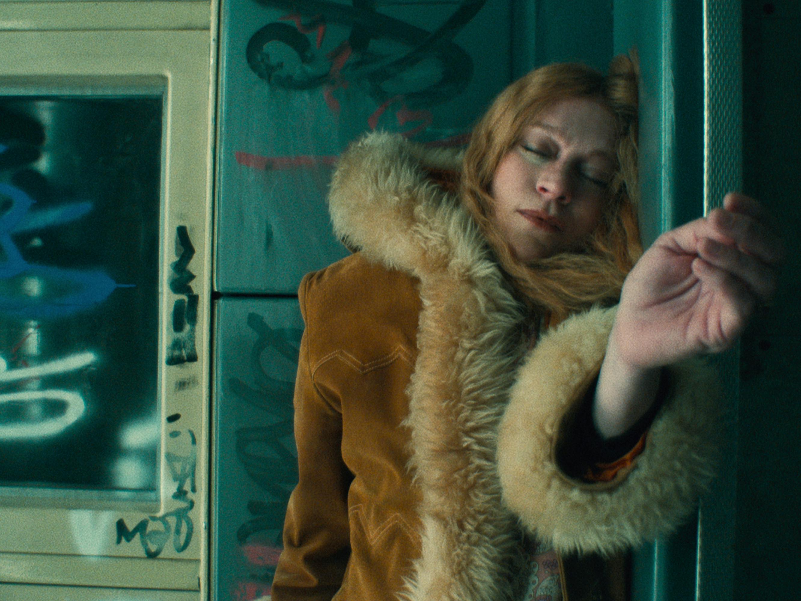 Nora (Chloë Sevigny) wears a fabulous fur trimmed coat and smokes a cigarette against a grafitted wall.