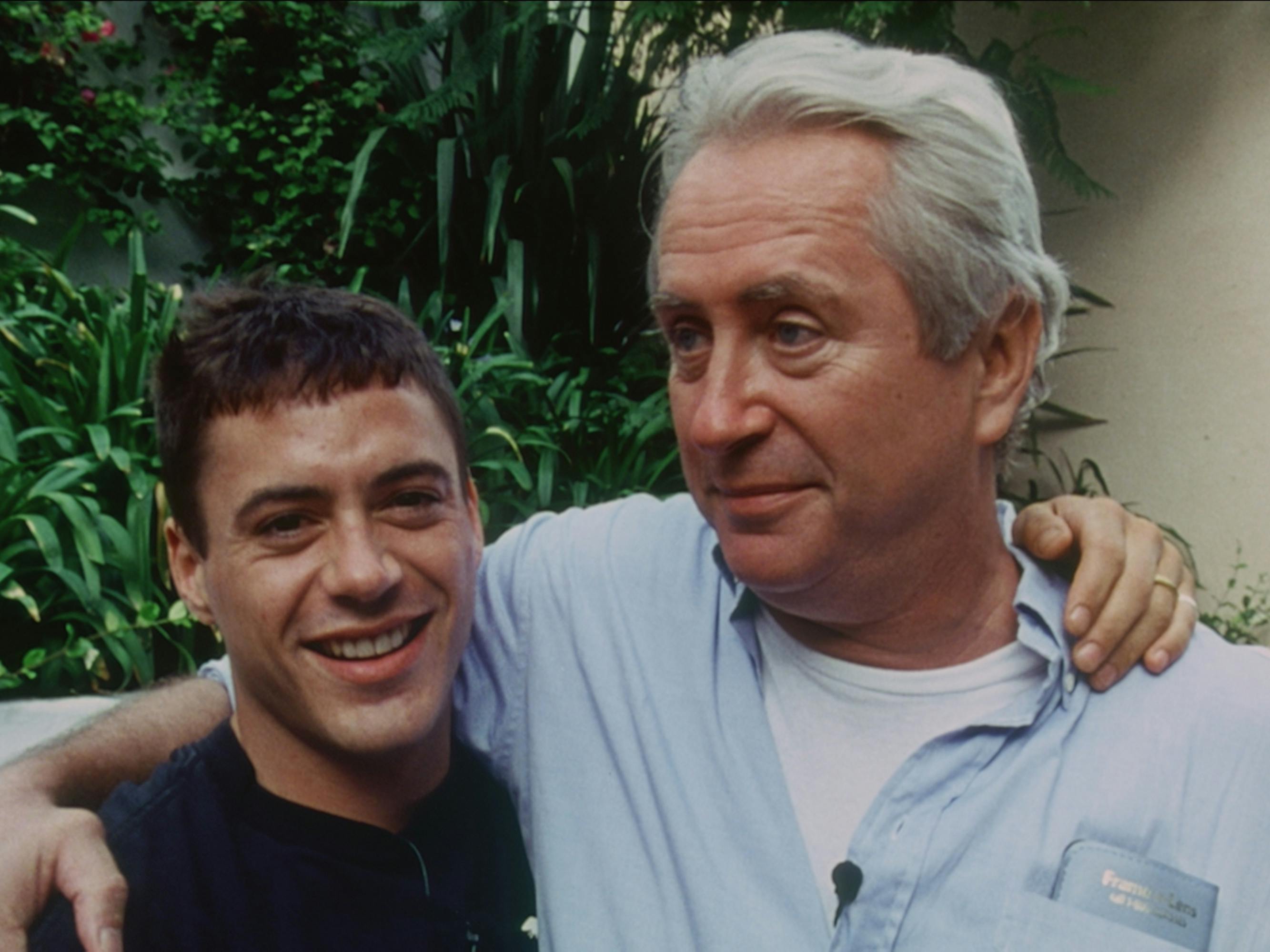 Robert Downey Jr. and Robert Downey Sr. stand together with their arms around each other. Jr. wears a dark shirt, Sr. wears a light blue long-sleeved over a white t-shirt.