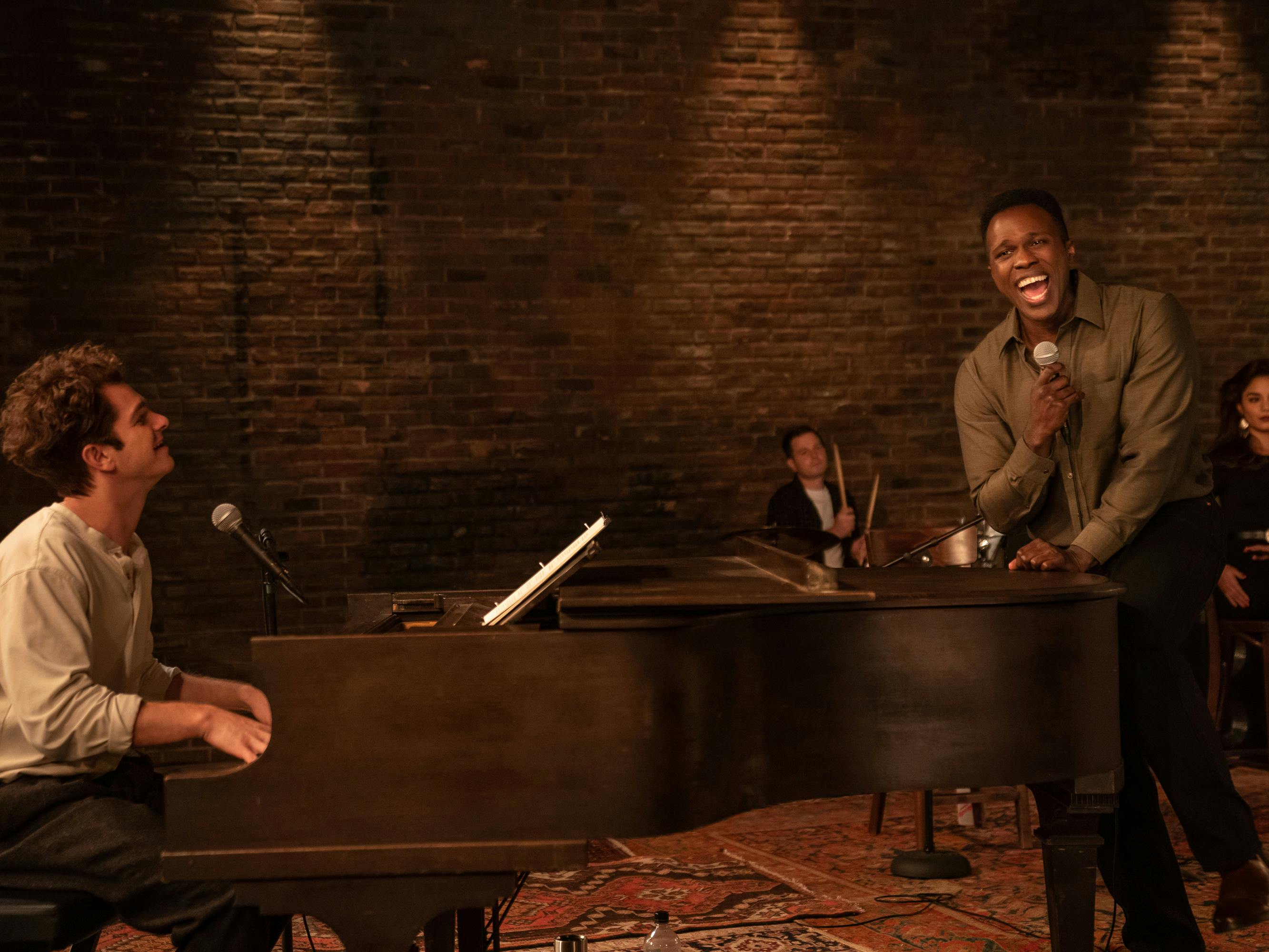 Andrew Garfield and Joshua Henry sing together around a piano into microphones. Behind them are the rest of the chorus and a brick wall.