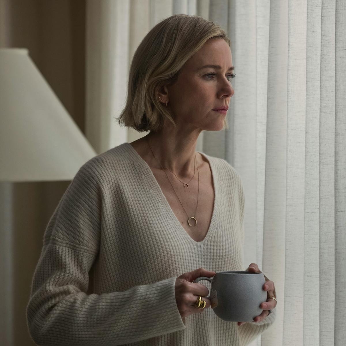 Nora Brannock (Naomi Watts) wears a beige sweater in her beige house and looks out the window, holding a mug.