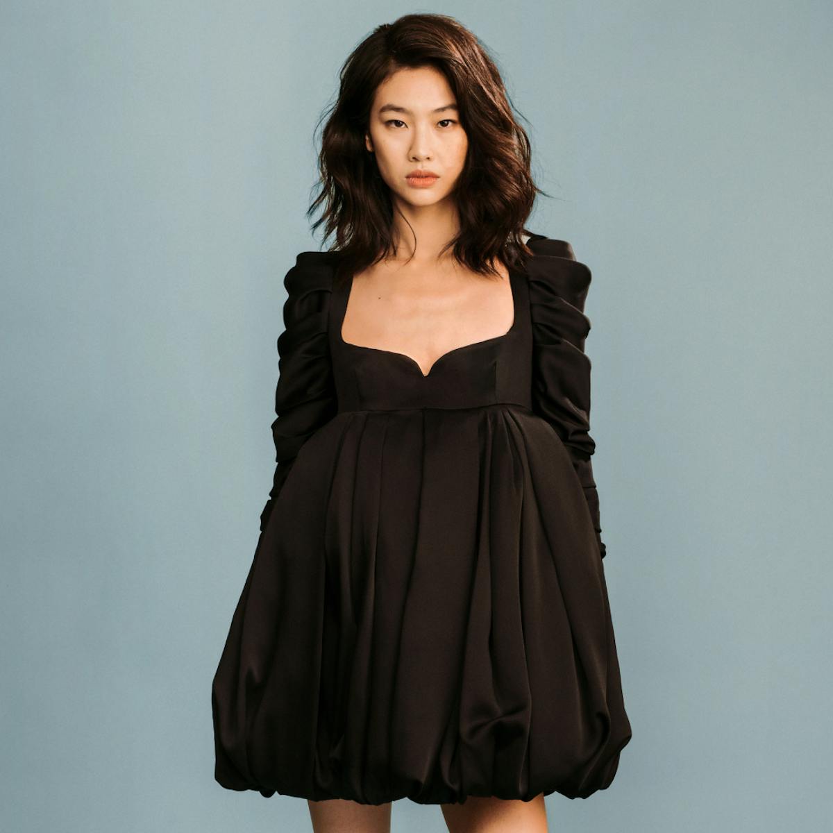 Jung Ho-yeon wears a black, short balloon dress and black boots, standing against a blue-gray background.
