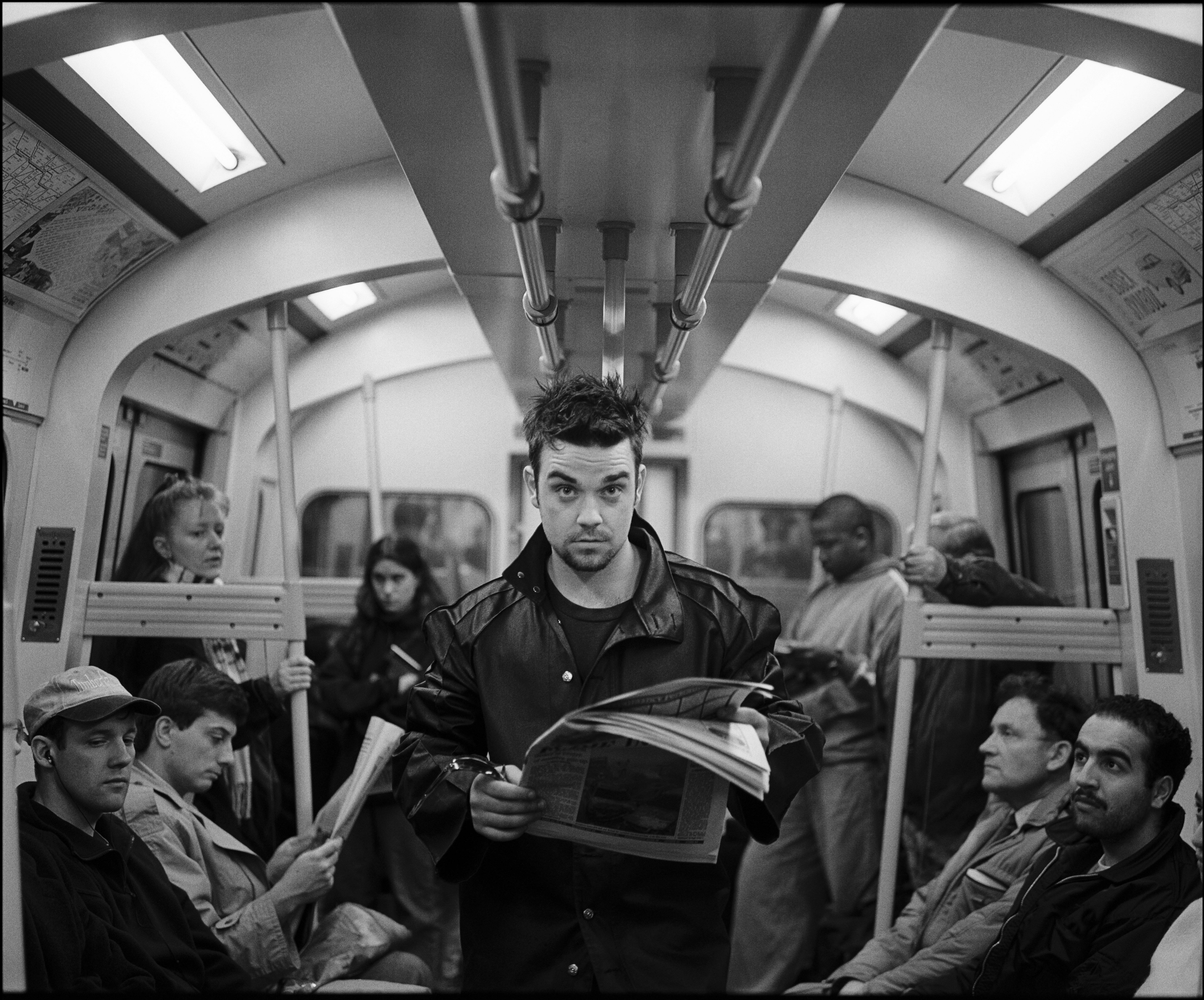 Robbie Williams stands on a subway reading a newspaper.