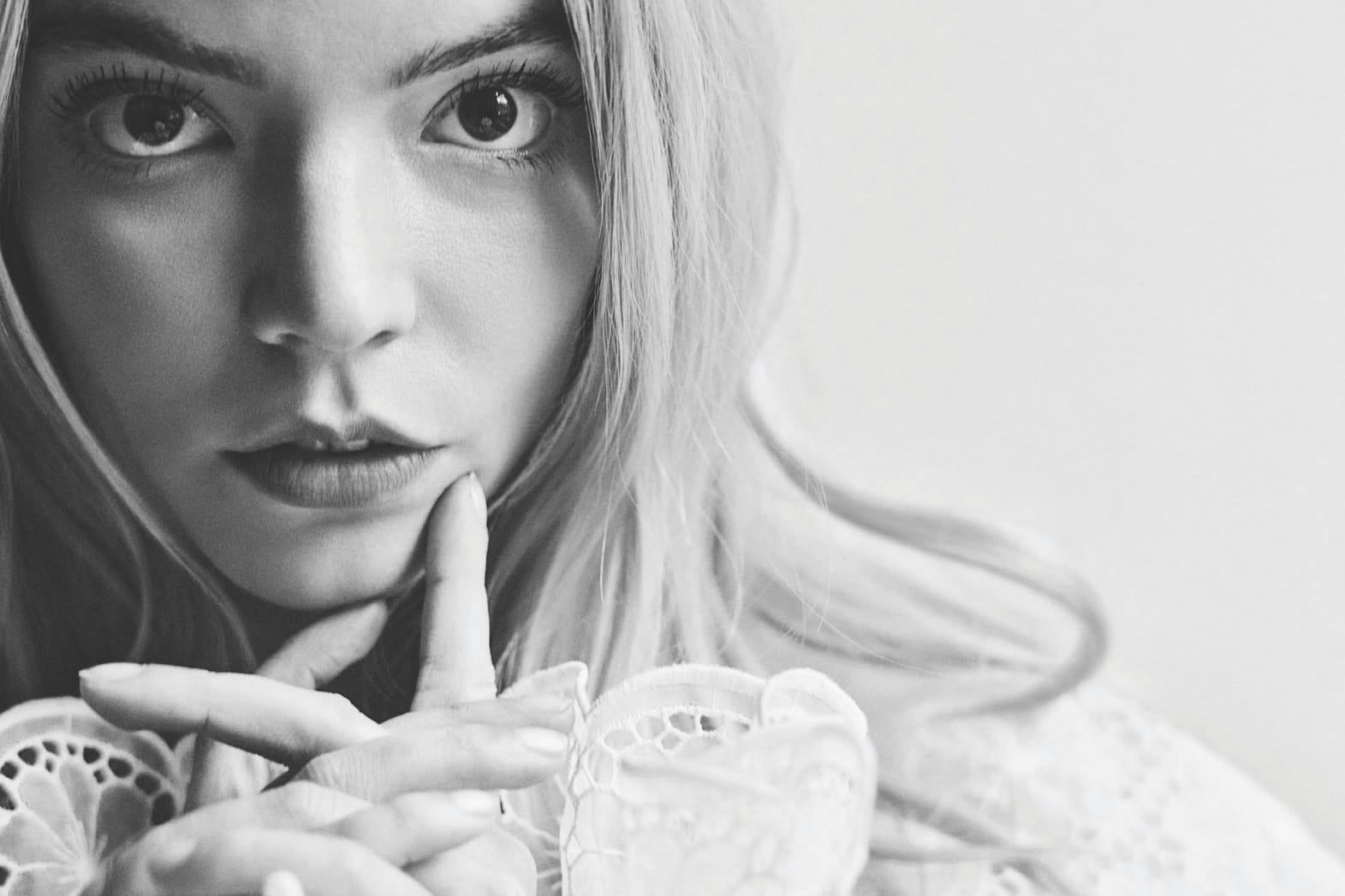 A black and white portrait of Anya Taylor-Joy. She wears all white and holds her finger against her cheek.