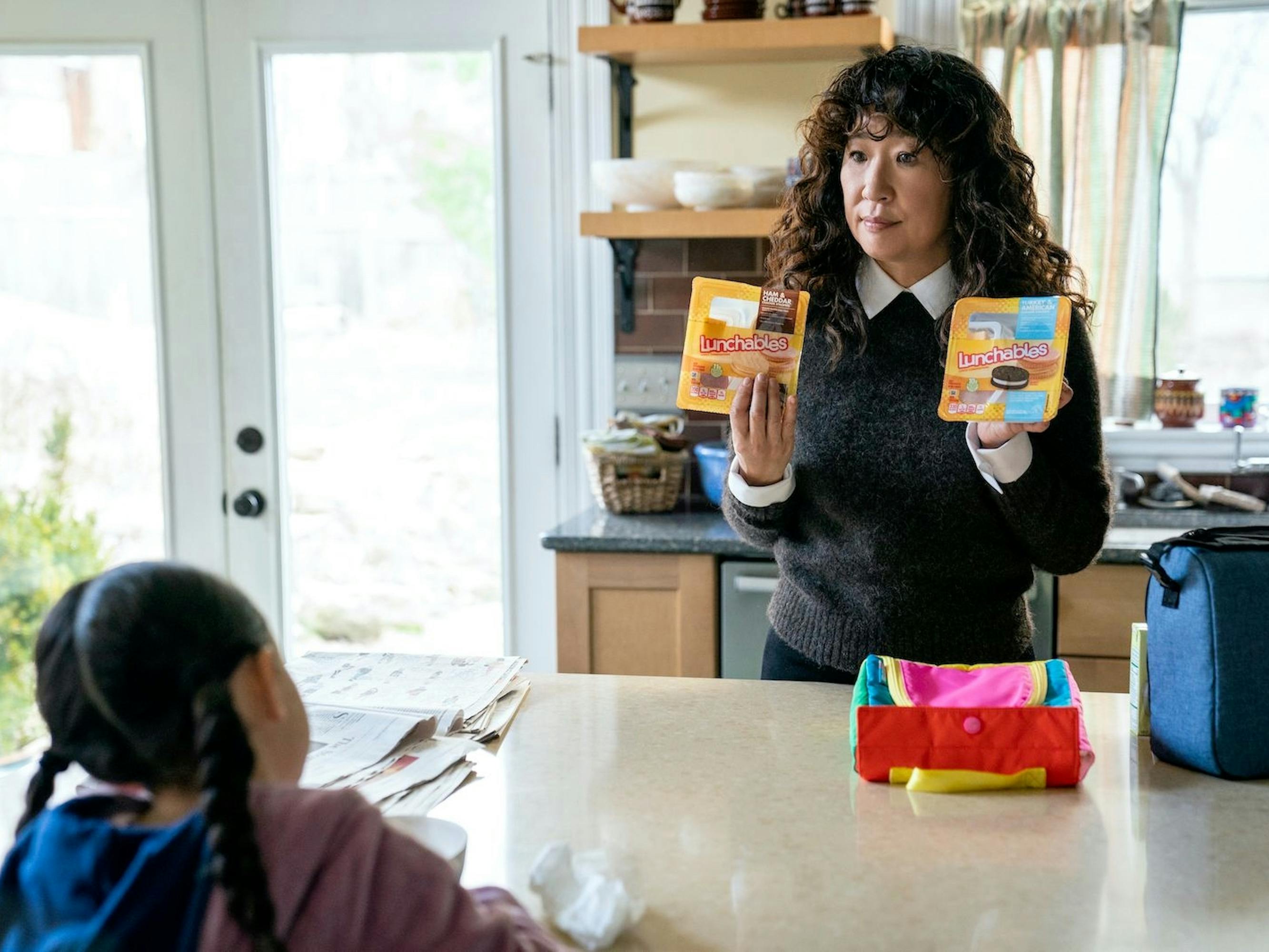 Ju-Hee (Everly Carganilla) and Dr. Ji-Yoon Kim (Sandra Oh) are in the kitchen together. Oh holds up two different types of lunchables for Carganilla to pick. The kitchen is flooded with light from the french doors.