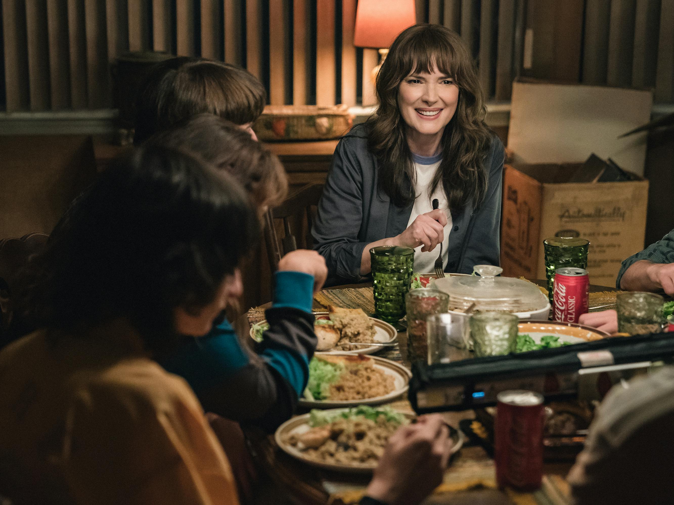 Mike Wheeler (Finn Wolfhard), Eleven (Millie Bobby Brown) Will Byers (Noah Schnapp), and Joyce Byers (Winona Ryder) sit around a full dinner table.