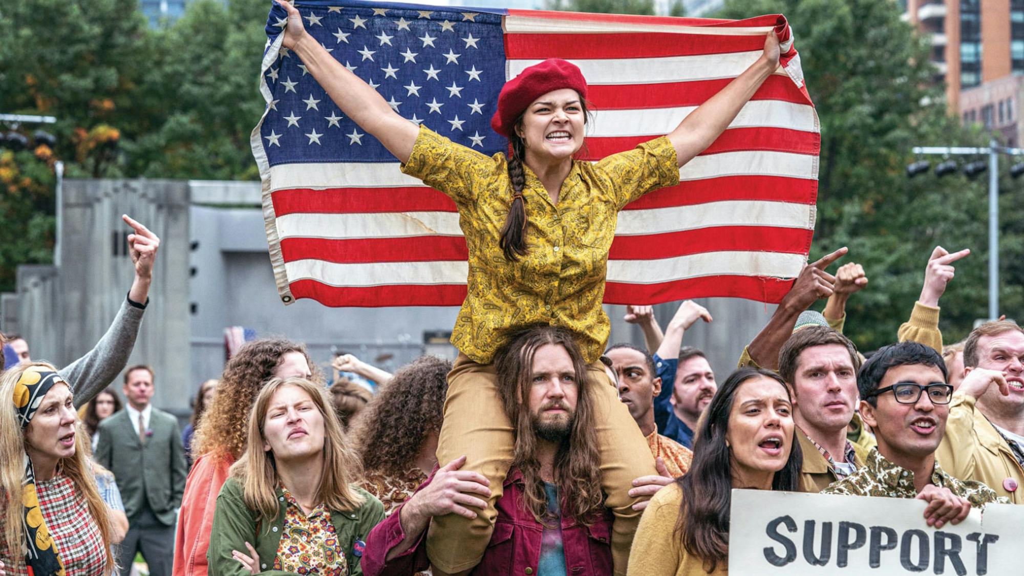 In this crowd scene, a young woman sits on a long-haired protester’s shoulders, wearing a red beret and holding an American flag outstretched. 