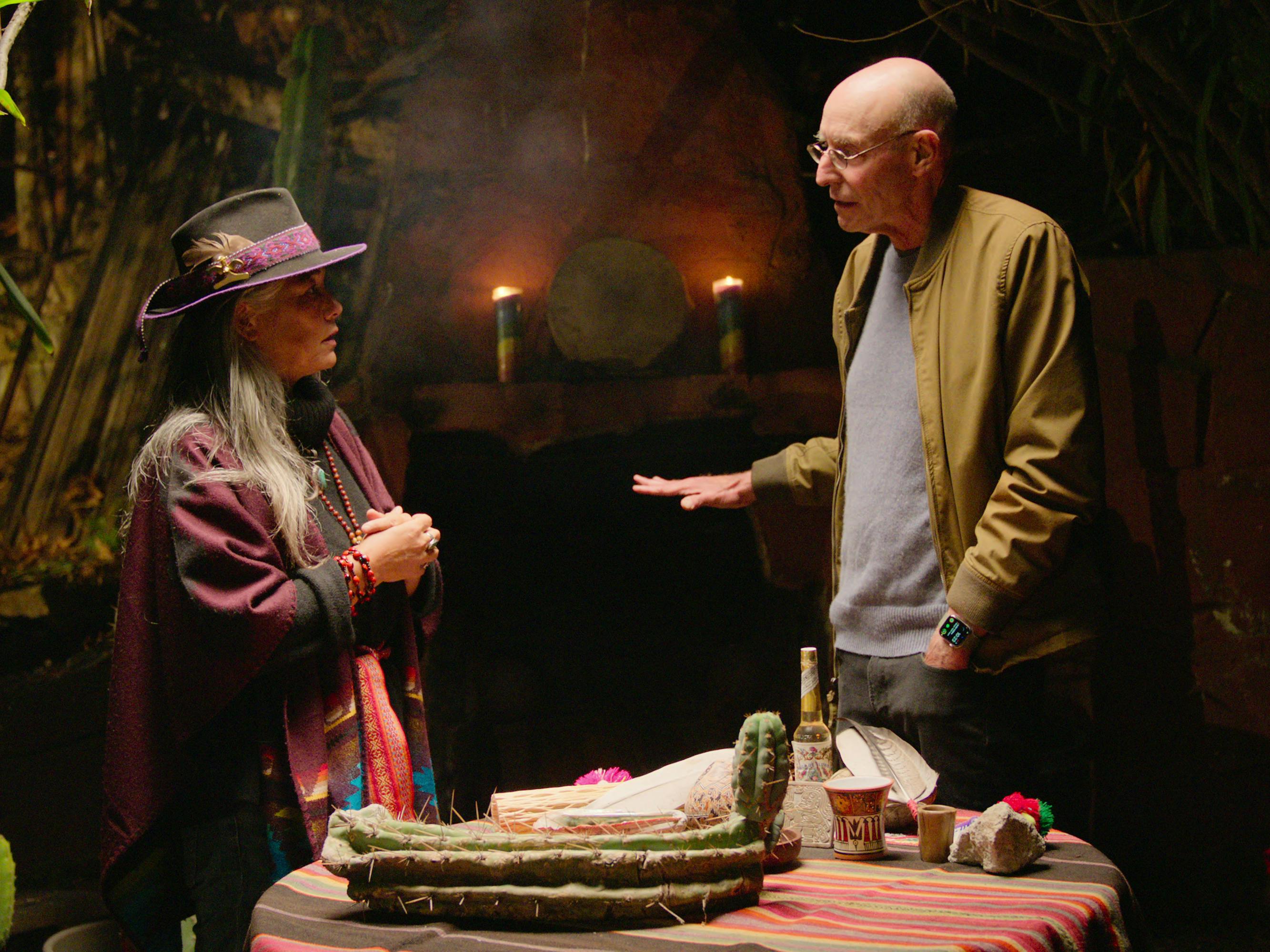 Erika Gagnon and Michael Pollan wearing a hat and red-maroon robes talk in a smoky dark room.