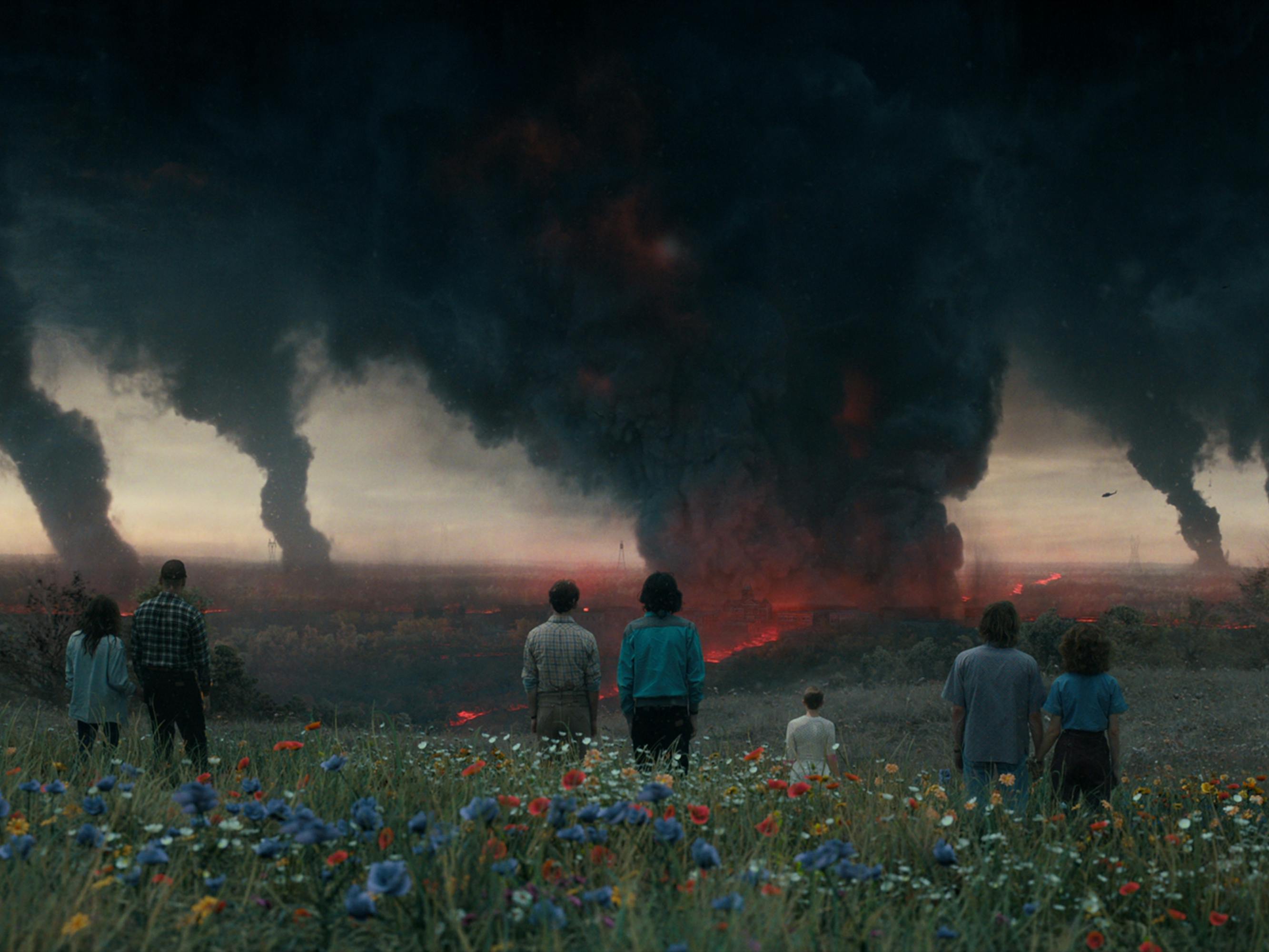 The characters of Stranger Things 4 look on at some explosions in a the middle of a flower-filled field. I hope their mullets don't get singed!