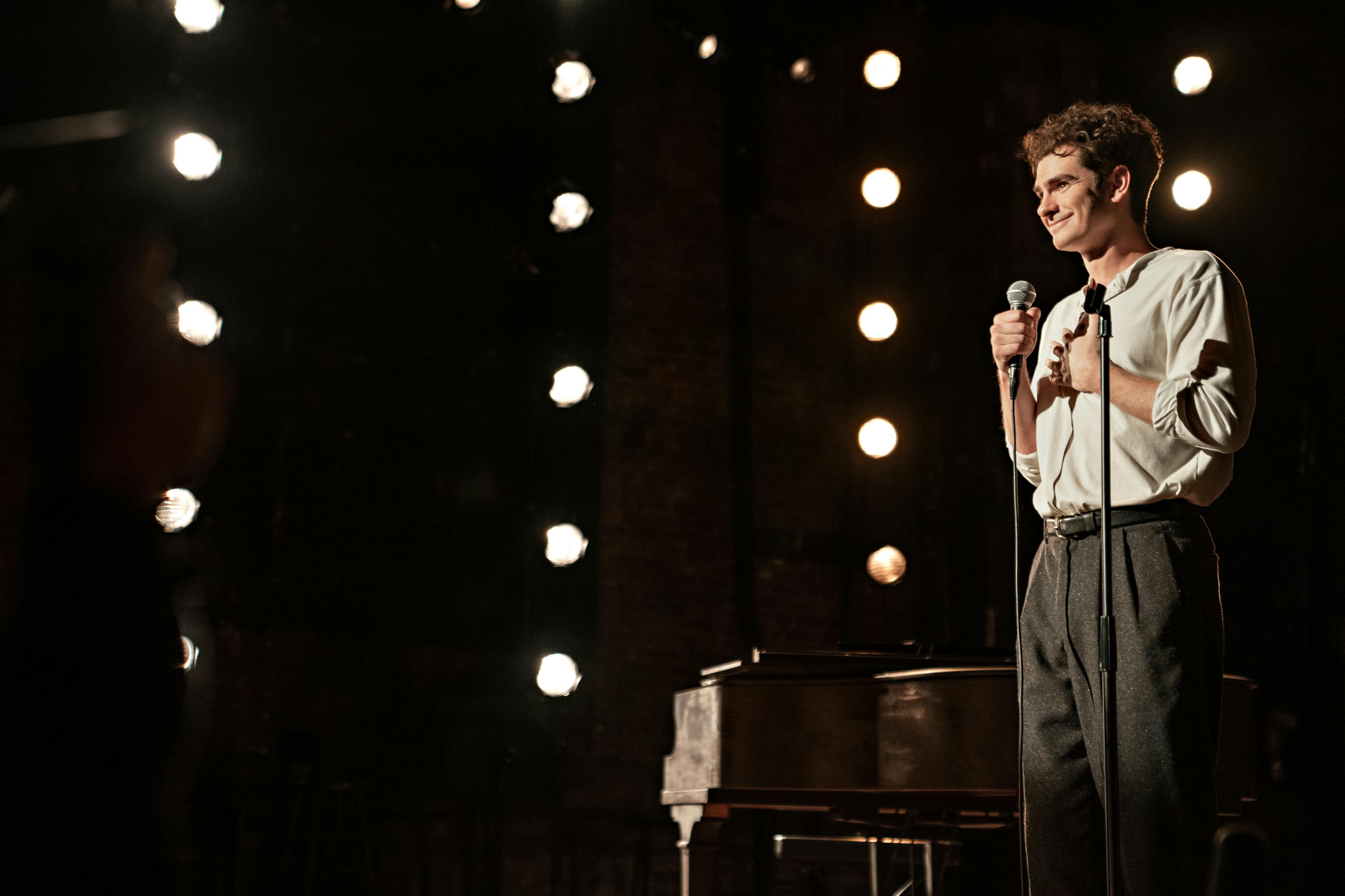 Andrew Garfield wears a white shirt and dark pants, and speaks into a microphone on a string-lit stage.