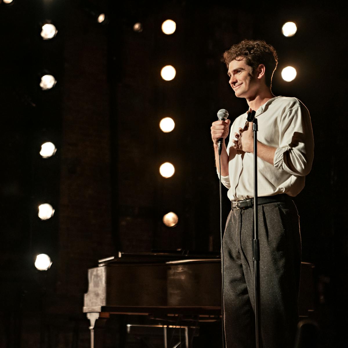 Andrew Garfield wears a white shirt and dark pants, and speaks into a microphone on a string-lit stage.