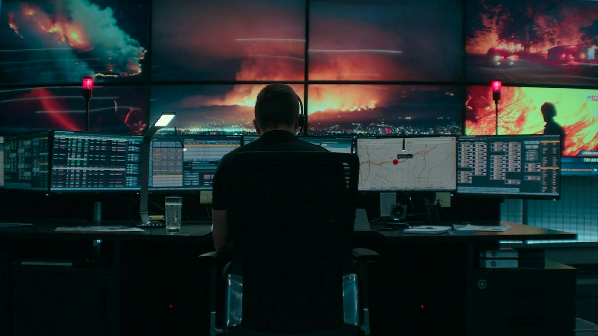 Joe Baylor wears a dark outfit and sits in front of a slew of desktop computers that show a fiery scene. 