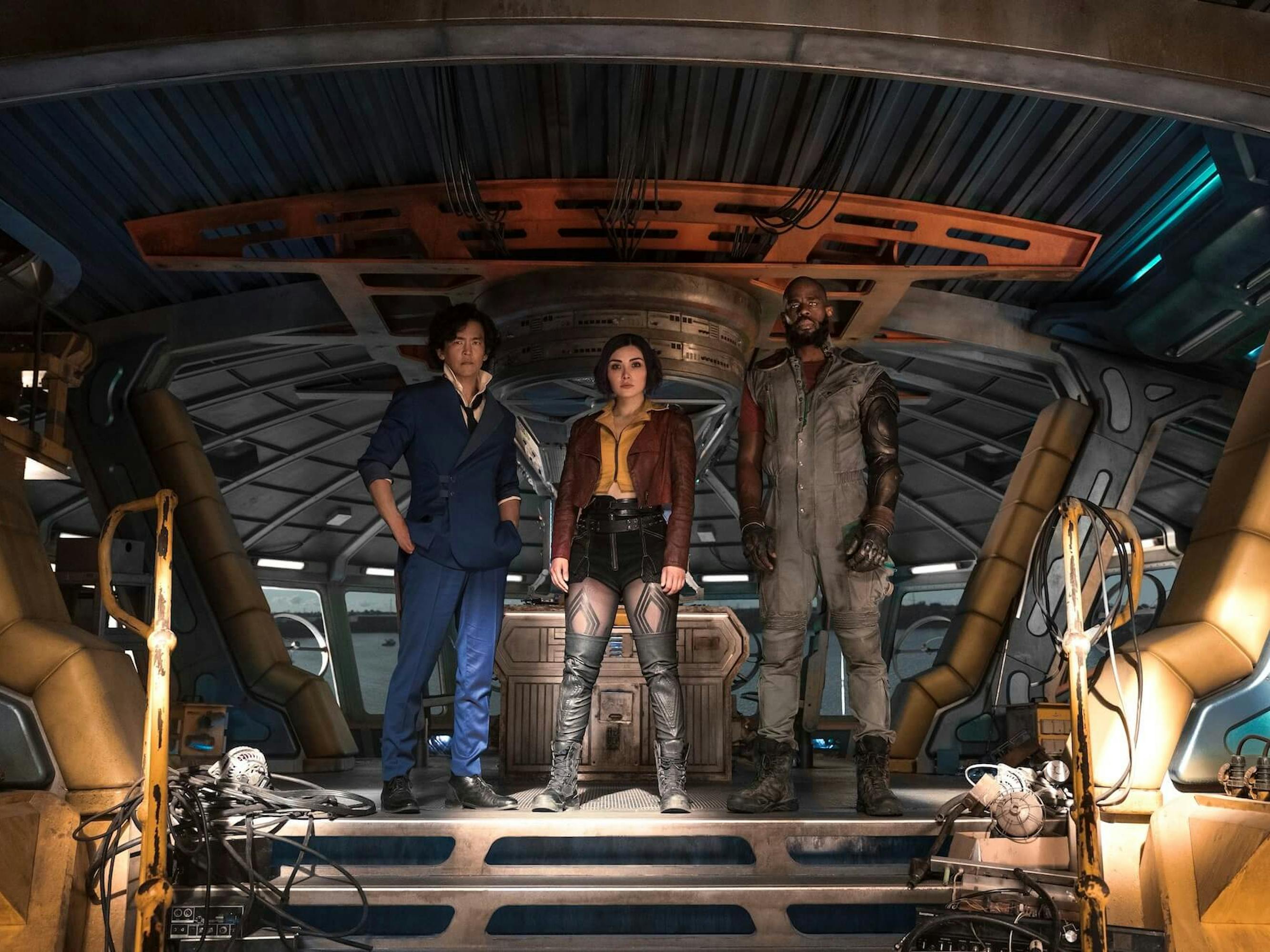 John Cho, Daniella Pineda, and Mustafa Shakir stand together. Cho wears a navy suit, Pineda wears a red jacket and leather boots, and Shakir wears a grey jumpsuit.
