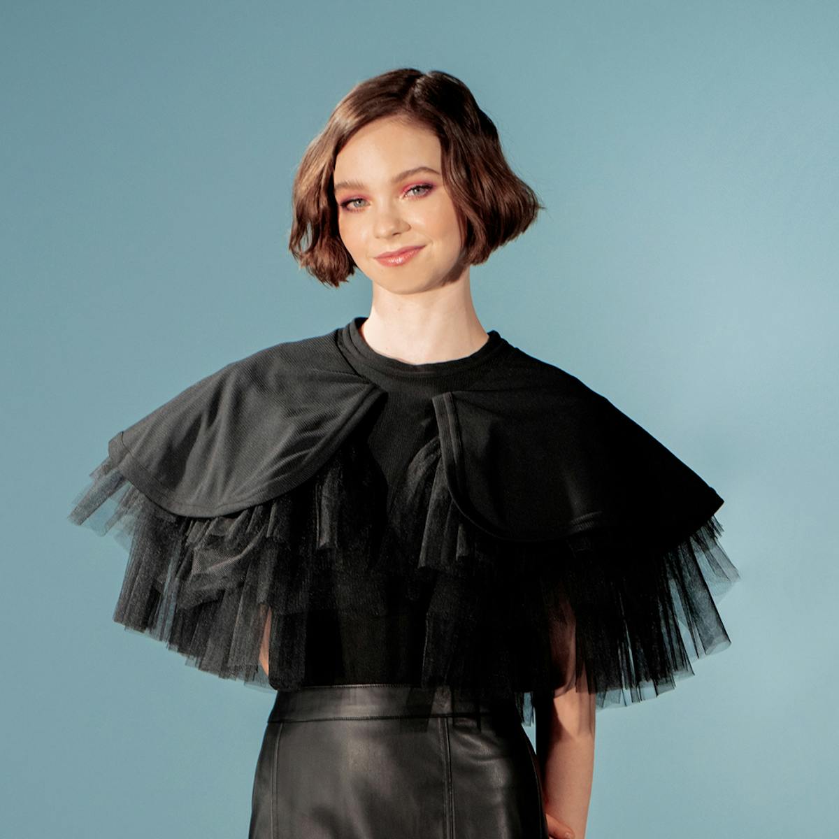 Emma Myers wears a black ensemble: a black leather skirt, a chiffon top with dignified sleeves, and strappy platforms. She stands on a black-and-white checkered floor against a blue background.
